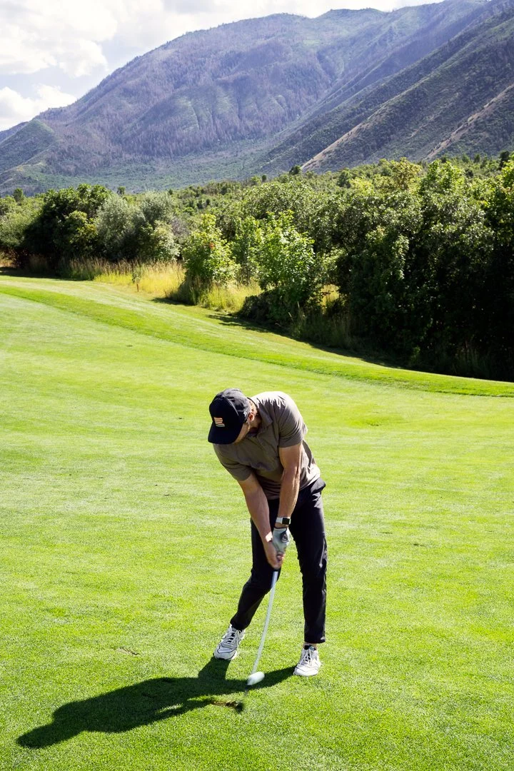 A person is preparing to hit a golf ball on a golf course with green grass, bushes, and mountains in the background on a sunny day.