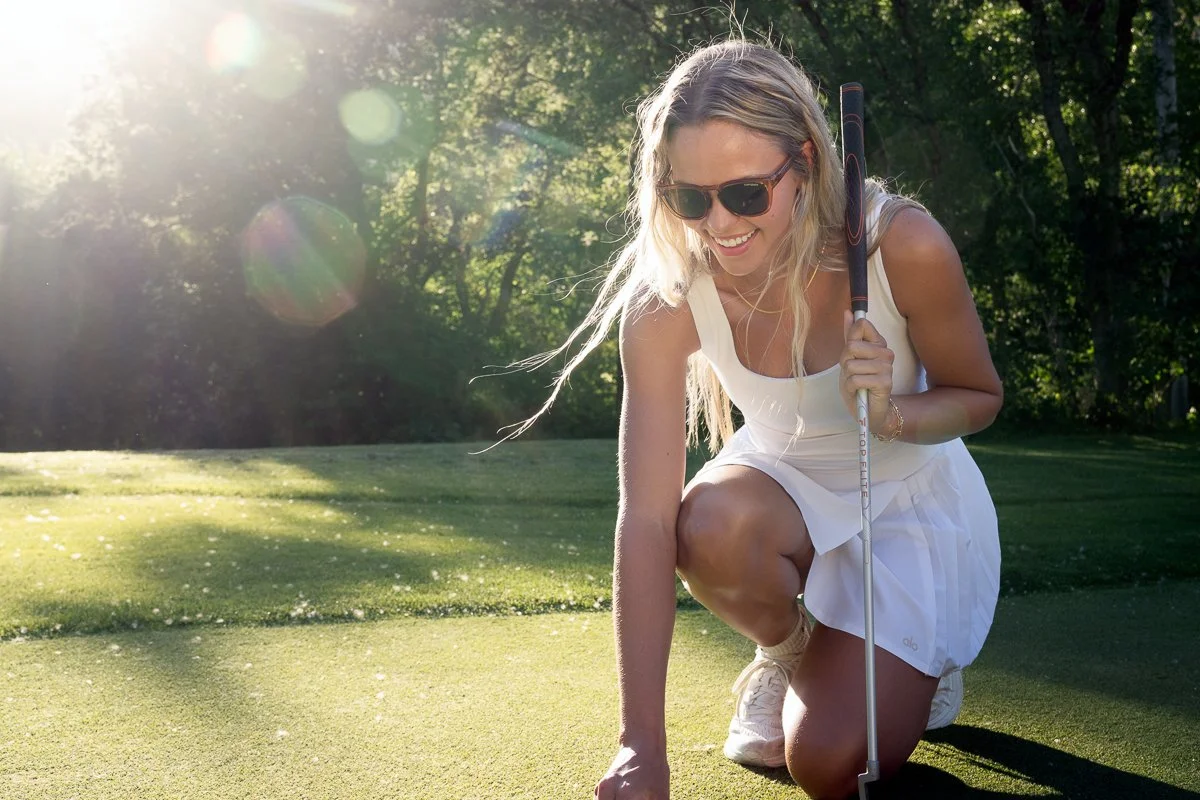 A woman wearing sunglasses, a white dress, and sneakers is smiling while kneeling on a golf course, holding a golf club, with sunlight filtering through trees in the background.