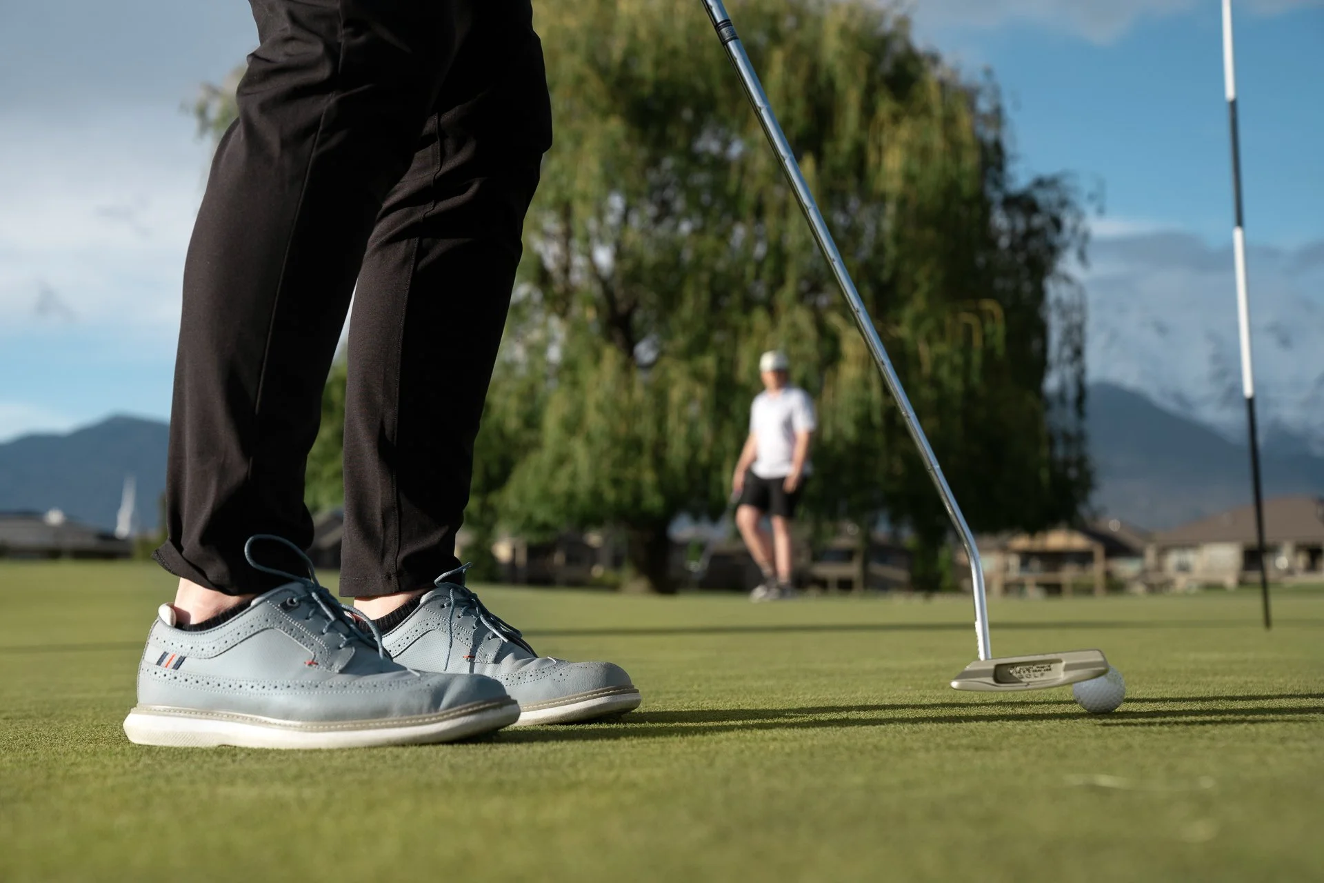Close-up of a golfer preparing to putt on the green with a golf club and ball, background shows another golfer, trees, houses, and mountains under a clear sky.