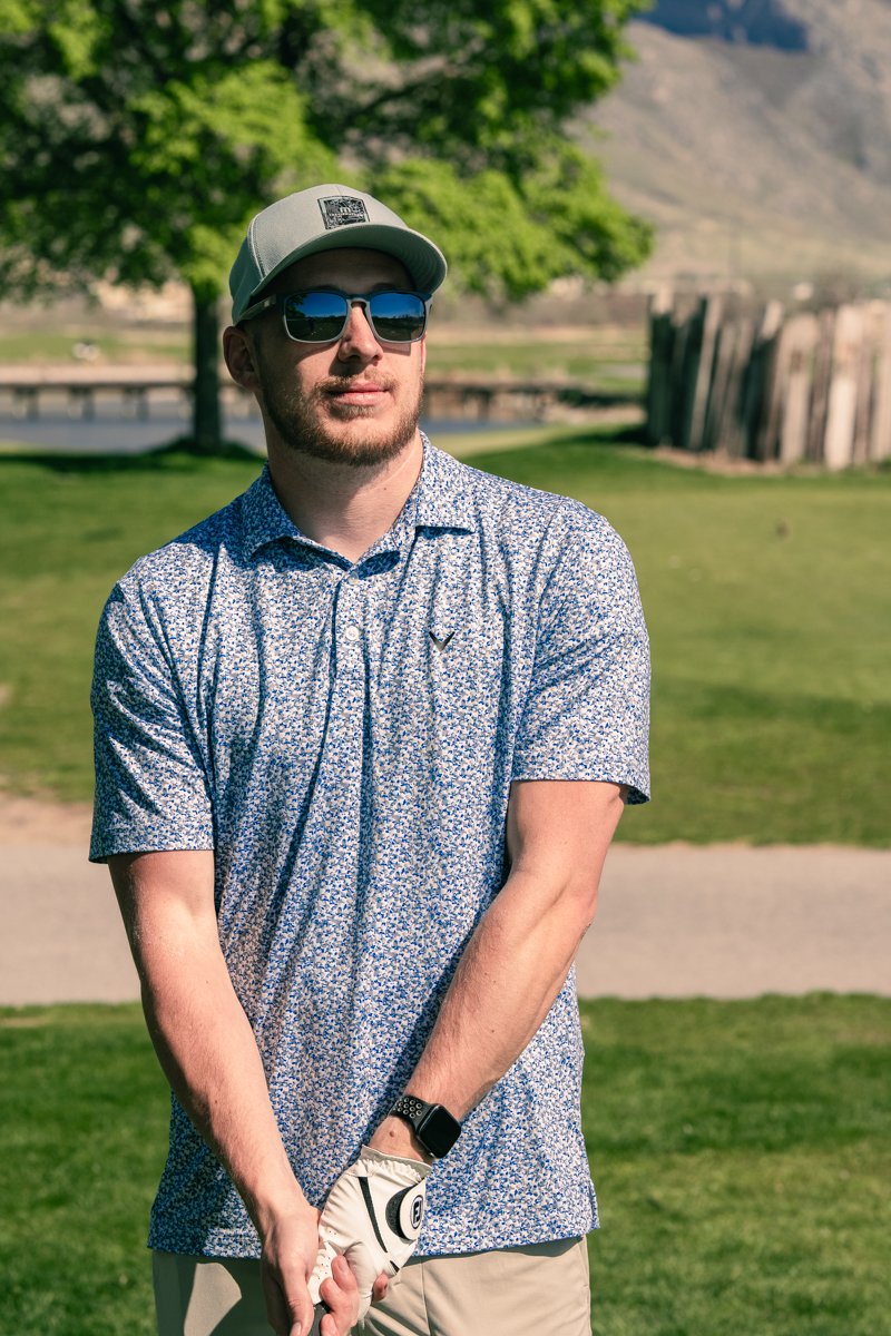 A man wearing sunglasses, a baseball cap, and a patterned polo shirt stands on a golf course holding a golf club with a golf glove. Background includes green grass, trees, and a fence.