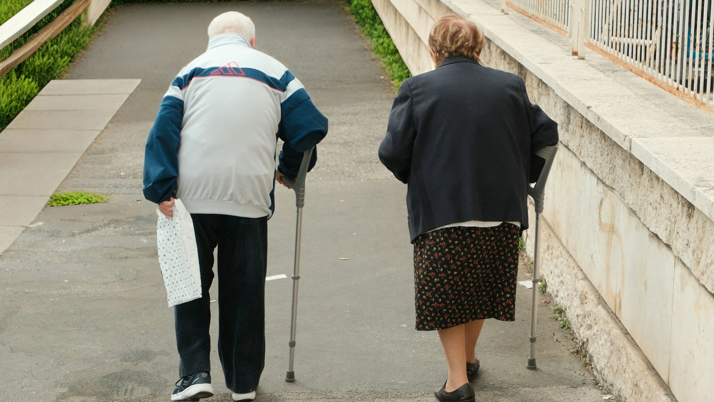 Two elderly women walking together, each using a cane, on a sidewalk next to a stone wall. One woman has short white hair and wears a light jacket with dark pants, while the other has curly brown hair and wears a dark jacket and floral skirt.