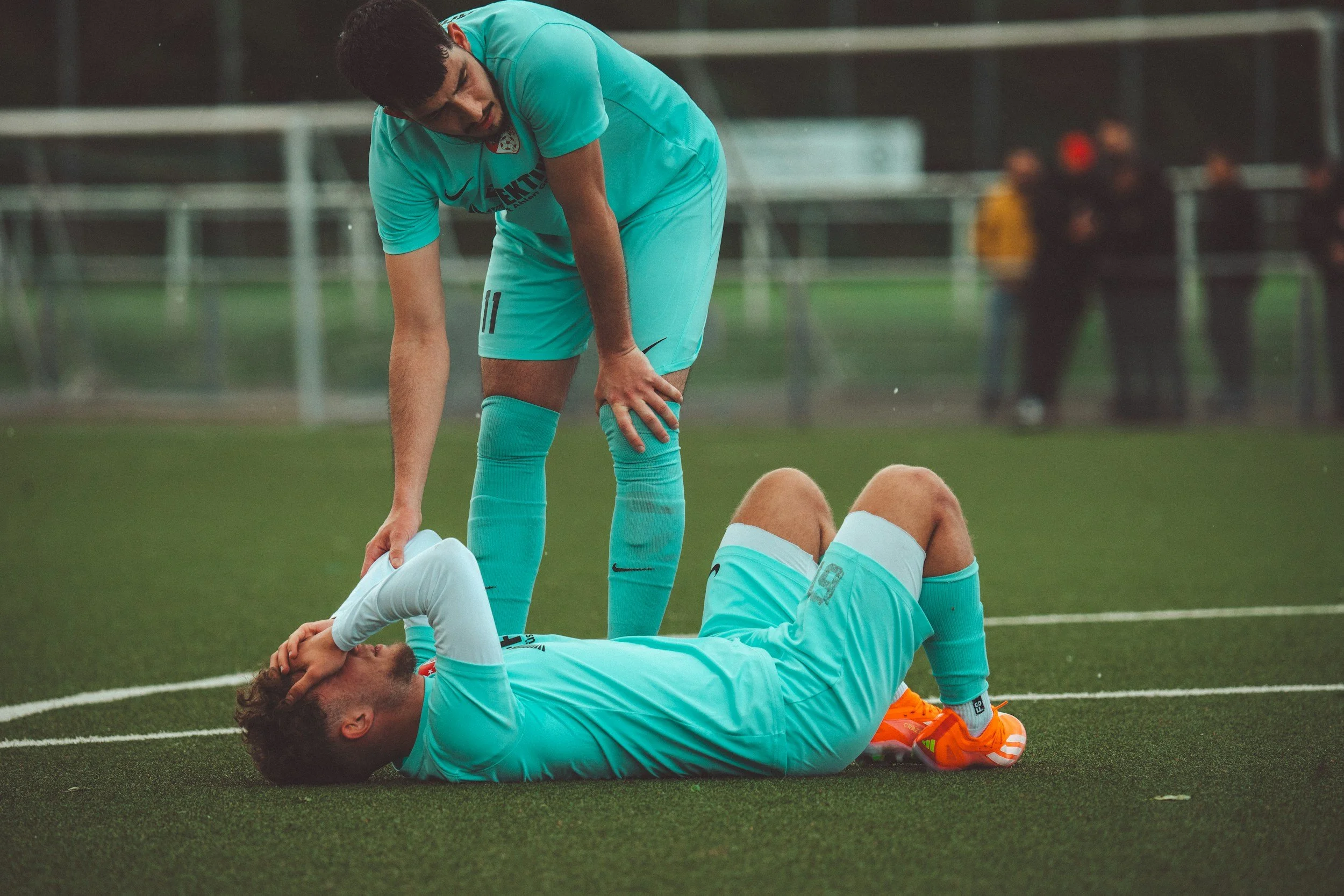 Soccer players in turquoise uniforms on the field; one player lying on the ground holding his head, another player standing over him. It appears to be a moment of injury or concern during a match.