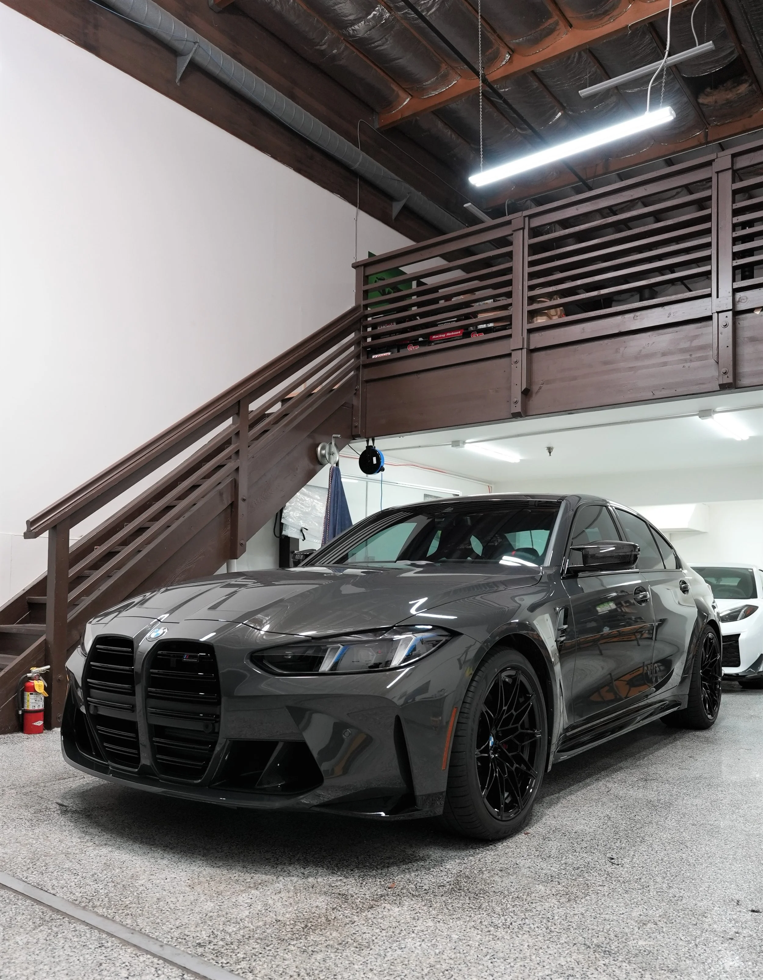 A gray BMW sports car parked inside a parking garage, with a black front grille, sleek headlights, and black rims. The garage has a textured gray floor, a wooden railing with a staircase, and exposed ceiling pipes and lighting.