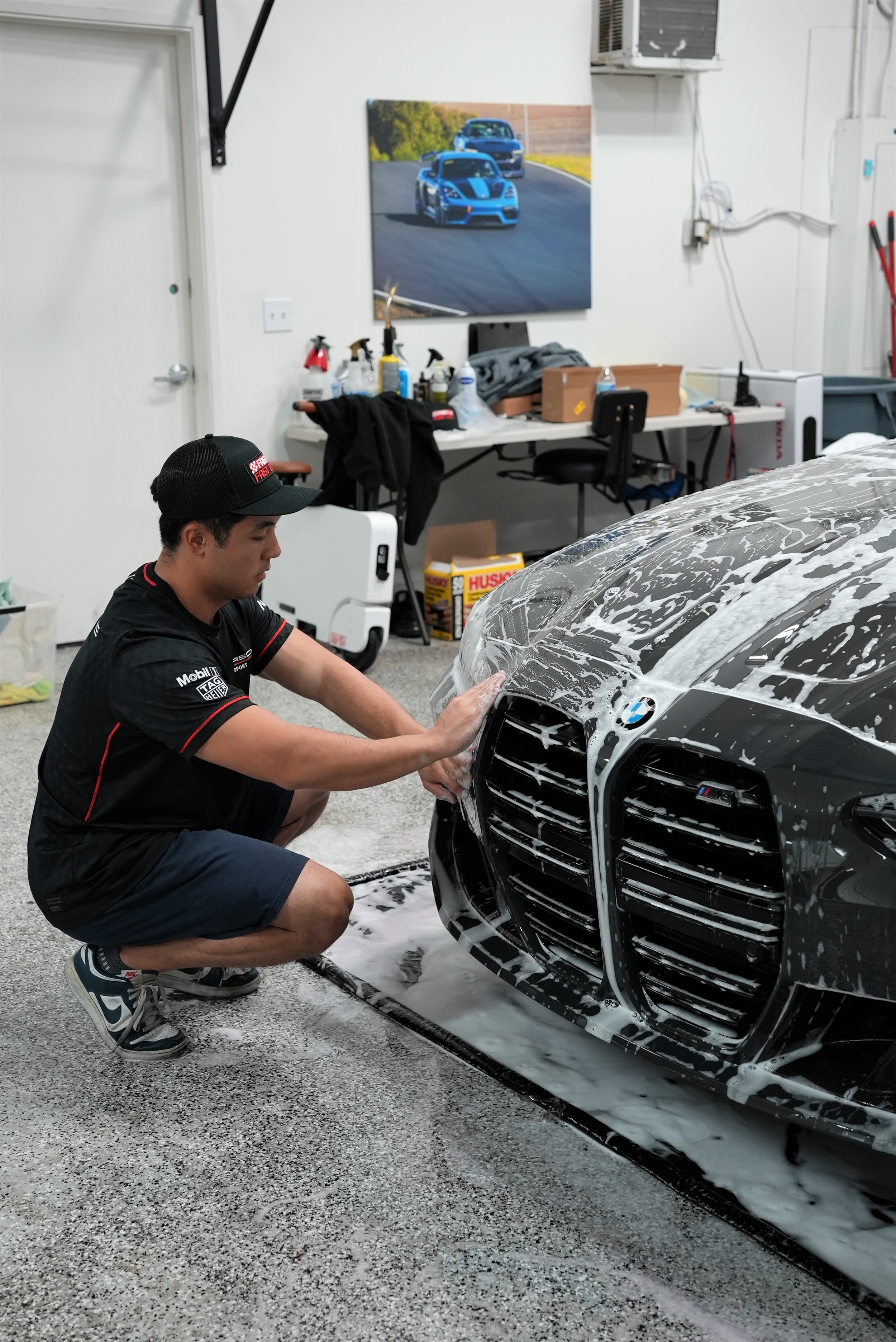 A man washing a black sports car with soap and water in a garage. The garage has a white wall, a table with cleaning supplies, and a picture of two blue cars racing on the wall.