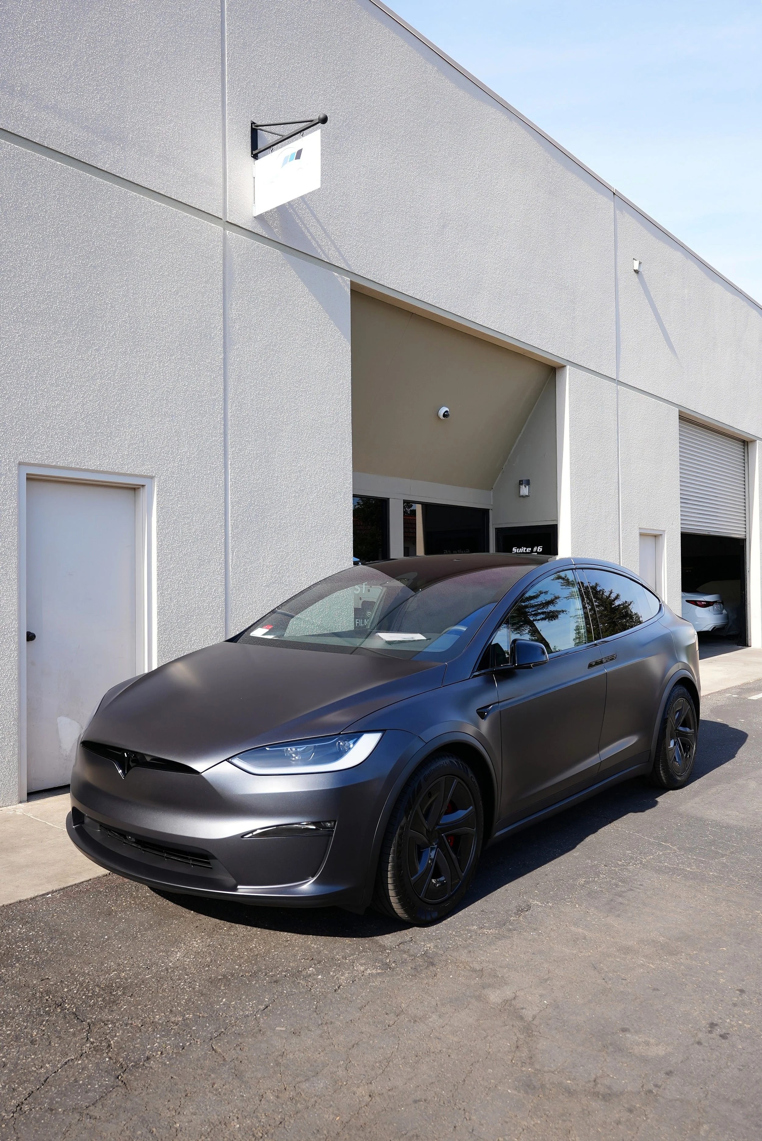 A black Tesla Model Y parked in front of a commercial building with a gray textured exterior wall and white doors.