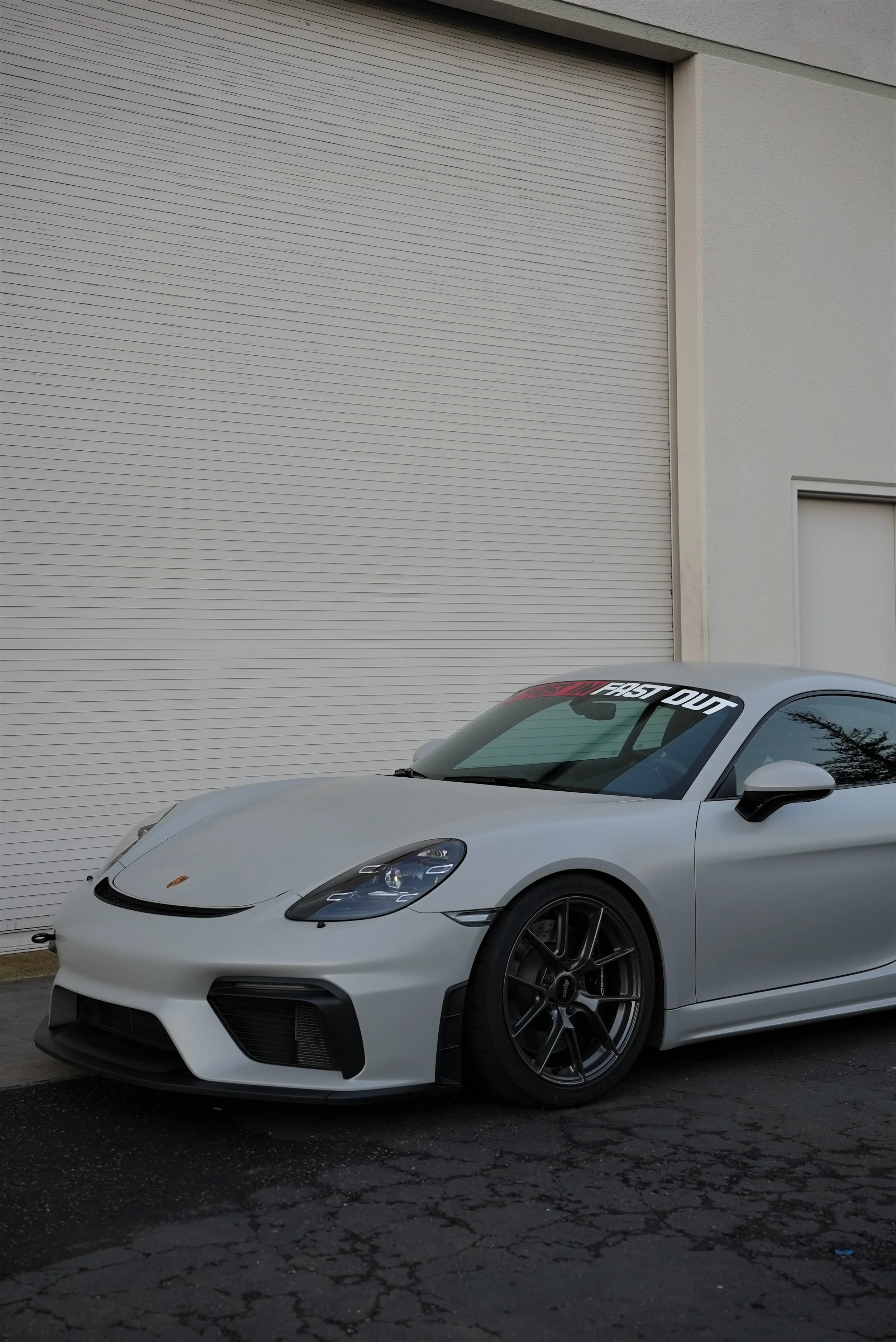 A silver sports car parked on a cracked asphalt surface in front of a beige building with a closed white roll-up garage door and a white door.