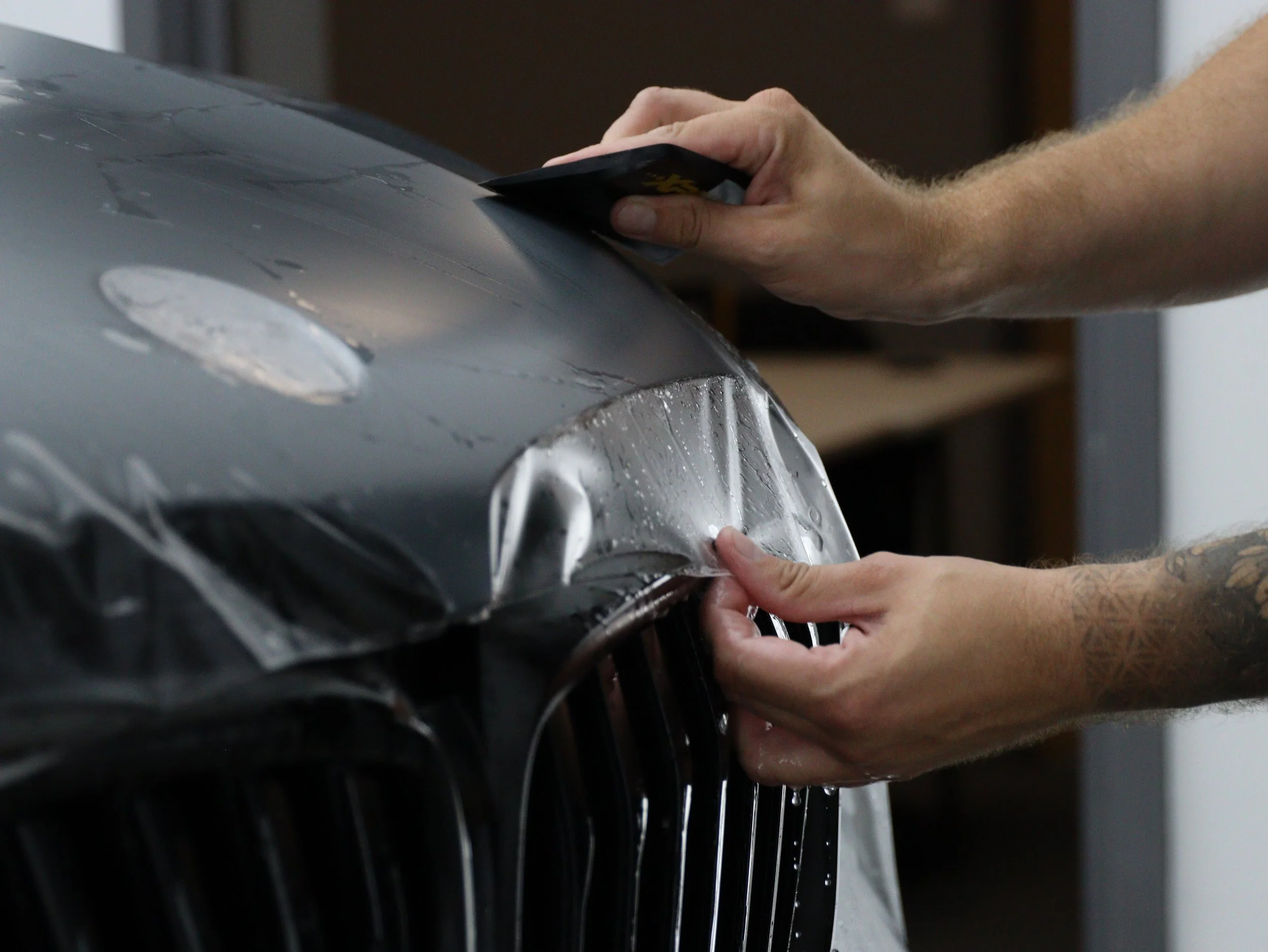 Hands applying clear film to a black car's front end, protecting the paint and grille.