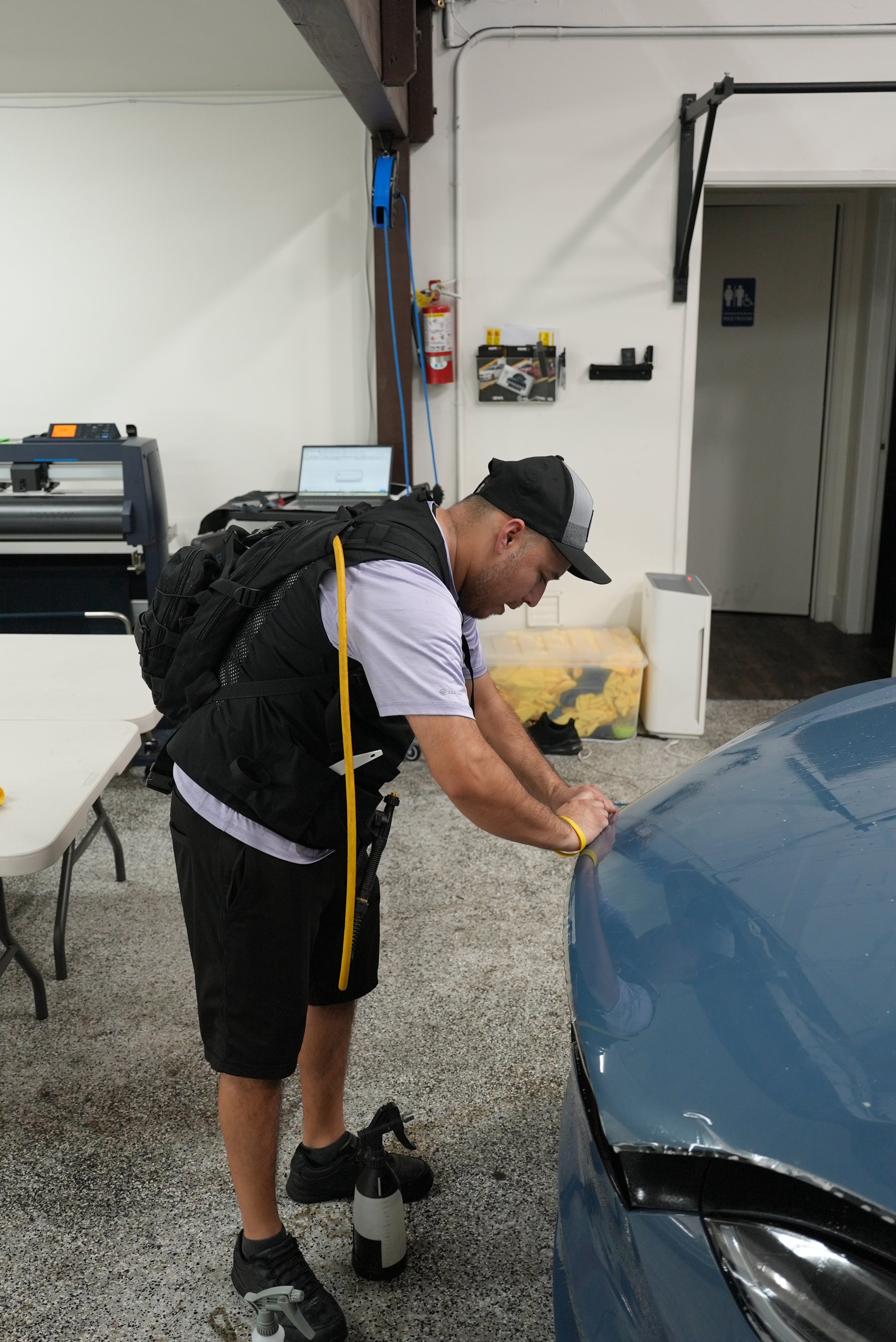 A man wearing a black and gray cap, black backpack, white t-shirt, black shorts, and black sneakers is polishing or detailing a blue car in a garage. The garage has a white wall with a fire extinguisher, some tools, a laptop, and other items.