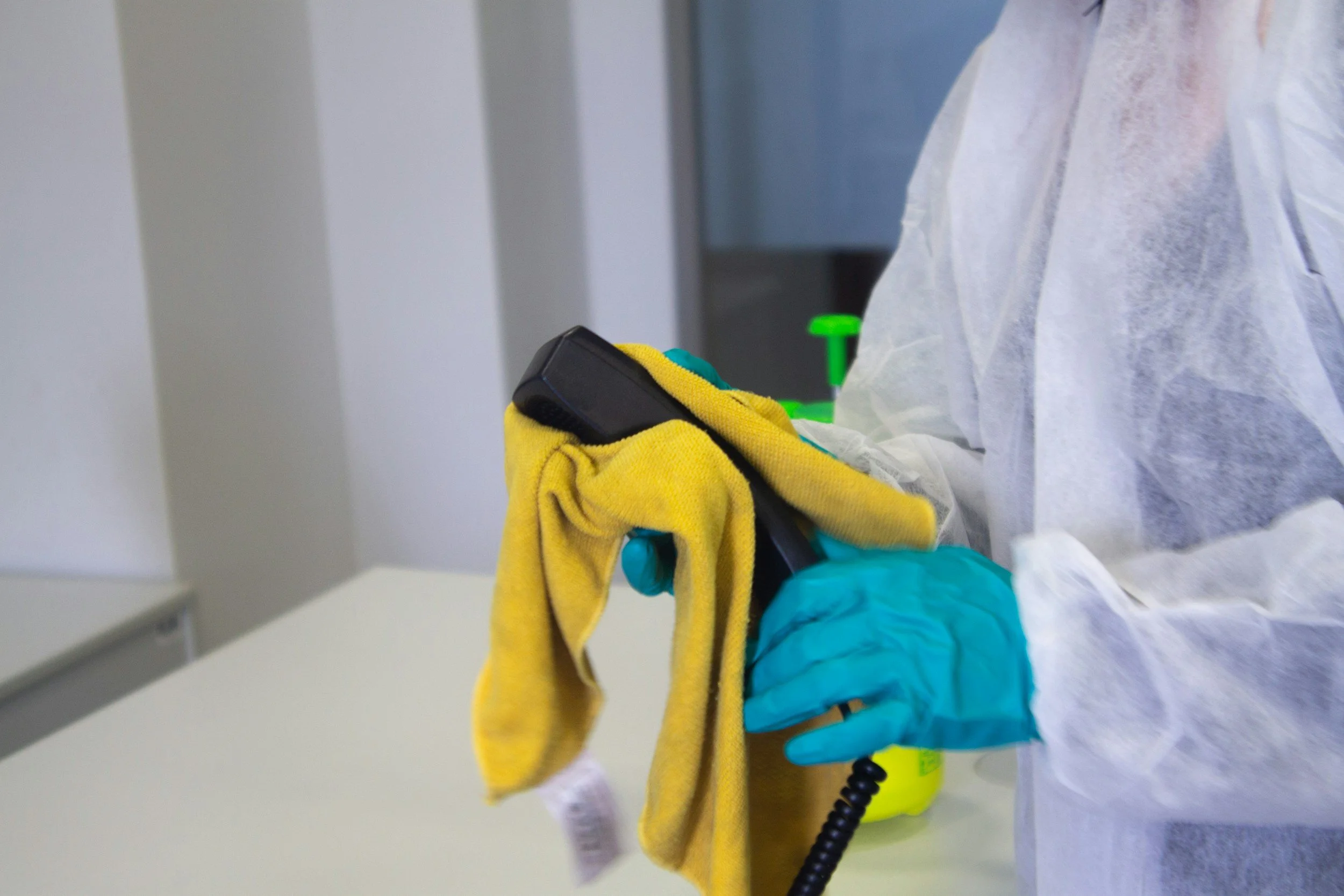 Person wearing protective gloves and gown cleaning a handheld device with a yellow cloth in a clinical setting.