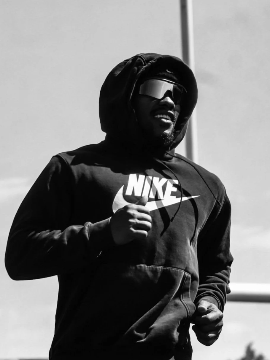 A man running outdoors, wearing a black Nike hoodie, sunglasses, and headphones, captured in black and white.
