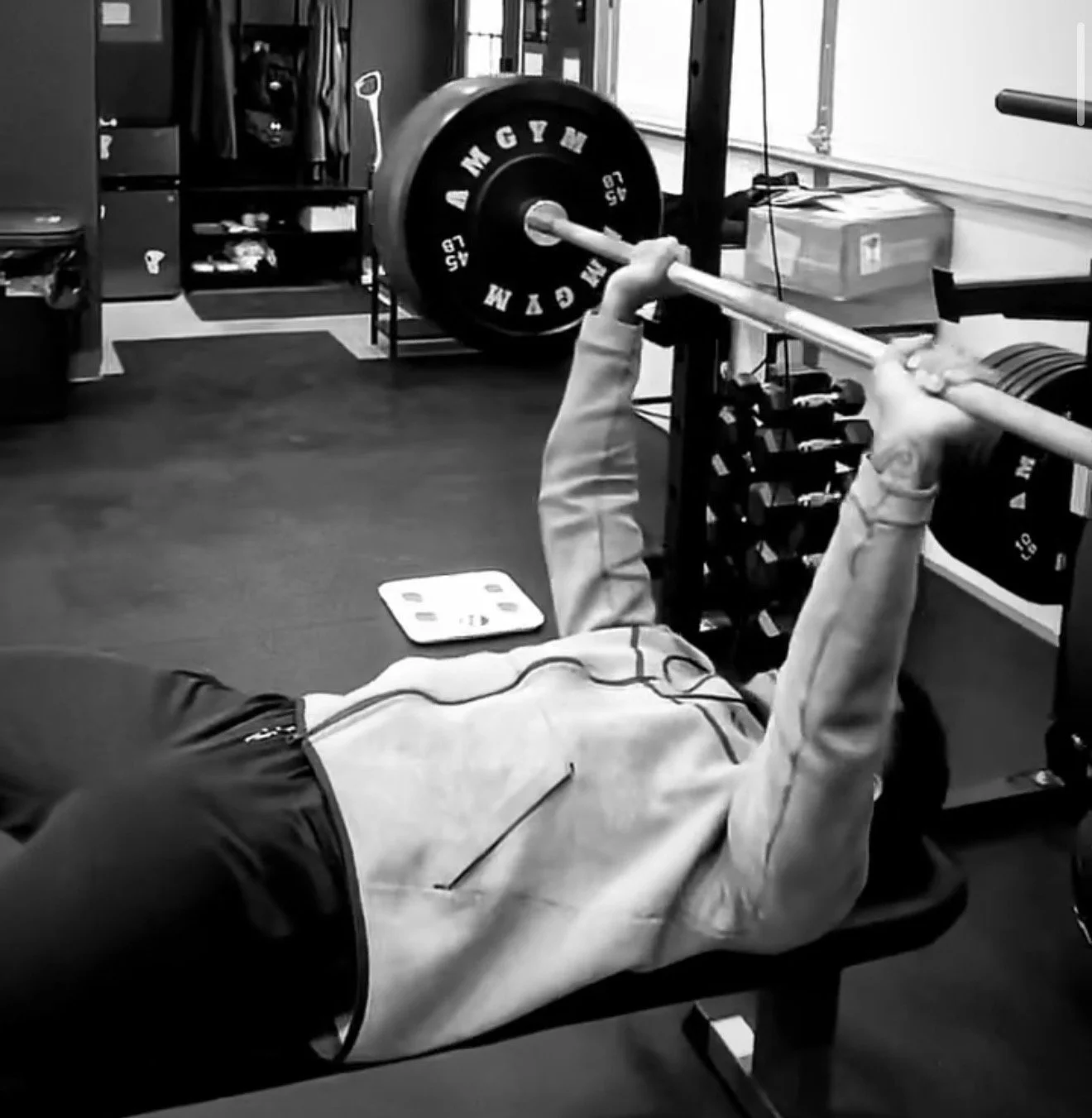A person lying on a workout bench lifting a barbell with weights in a gym, black and white photo.
