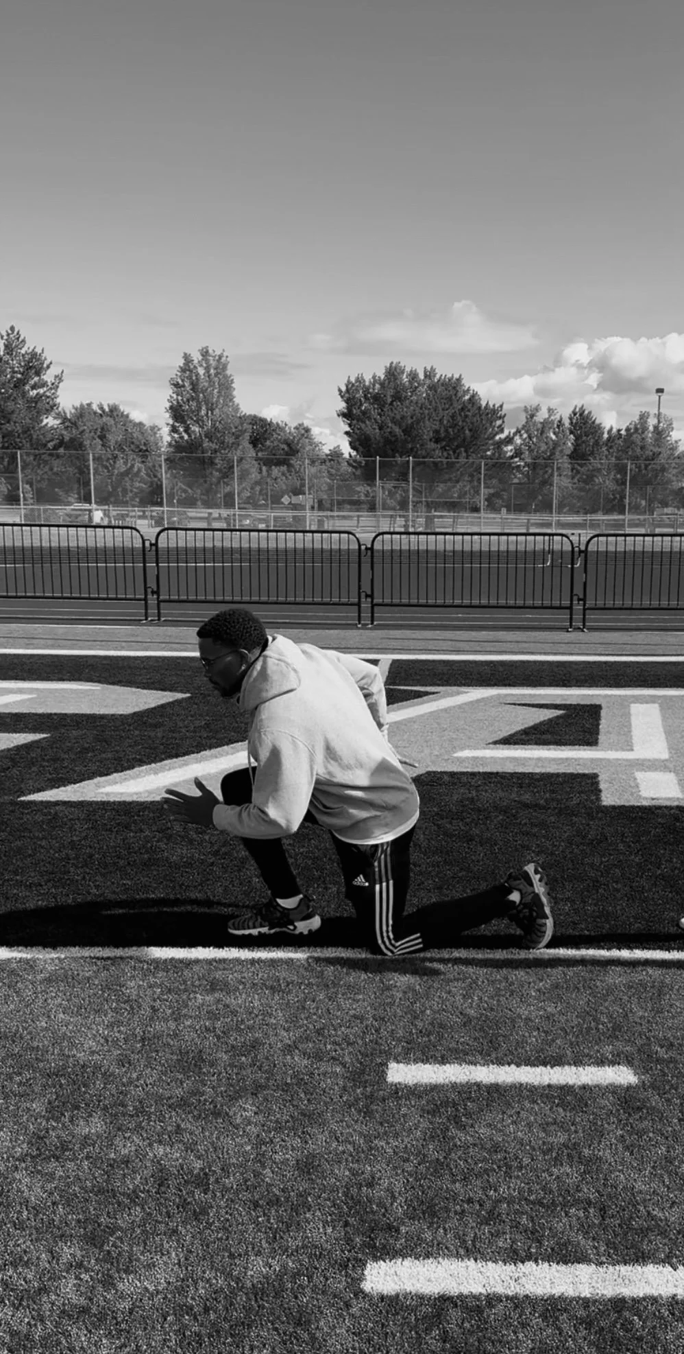 A person in athletic clothing kneeling on a sports field at sunset, with trees and a fence in the background, captured in black and white.