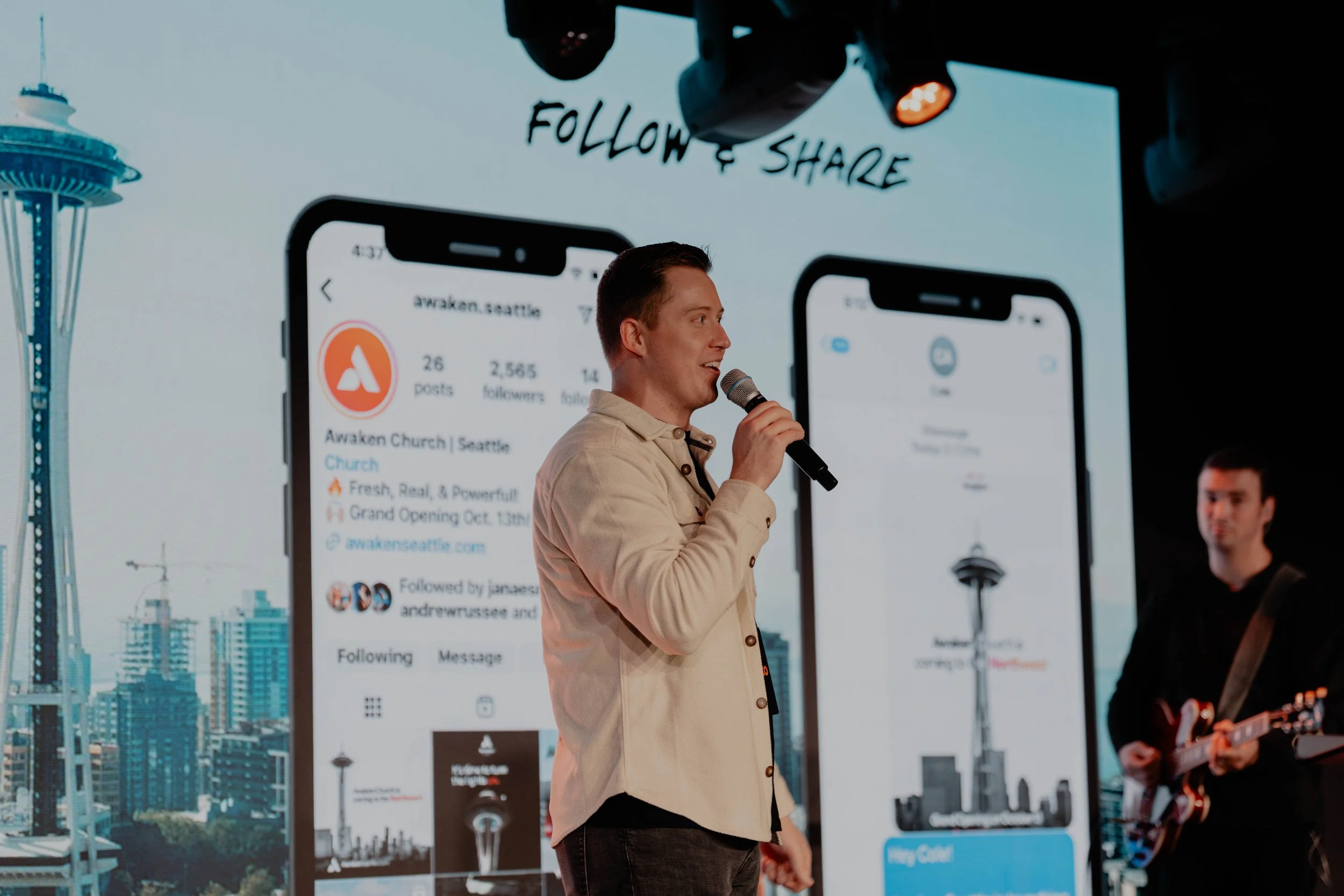 A man holding a microphone speaking on stage with a large screen behind him displaying social media profiles and images of the Seattle skyline, including the Space Needle.