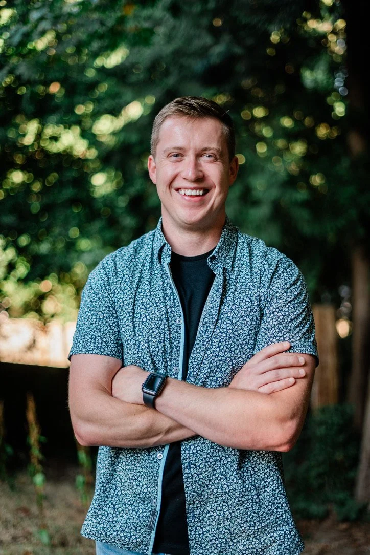 Young man smiling outdoors with arms crossed, wearing a patterned short-sleeve shirt and a smartwatch, standing in front of greenery.