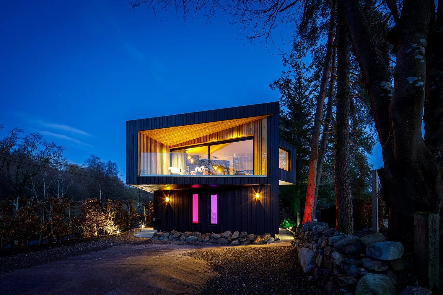 Loch Venachar Lodges. A modern lochside self catering house with blue skies at night. It is lit up with cool lighting.