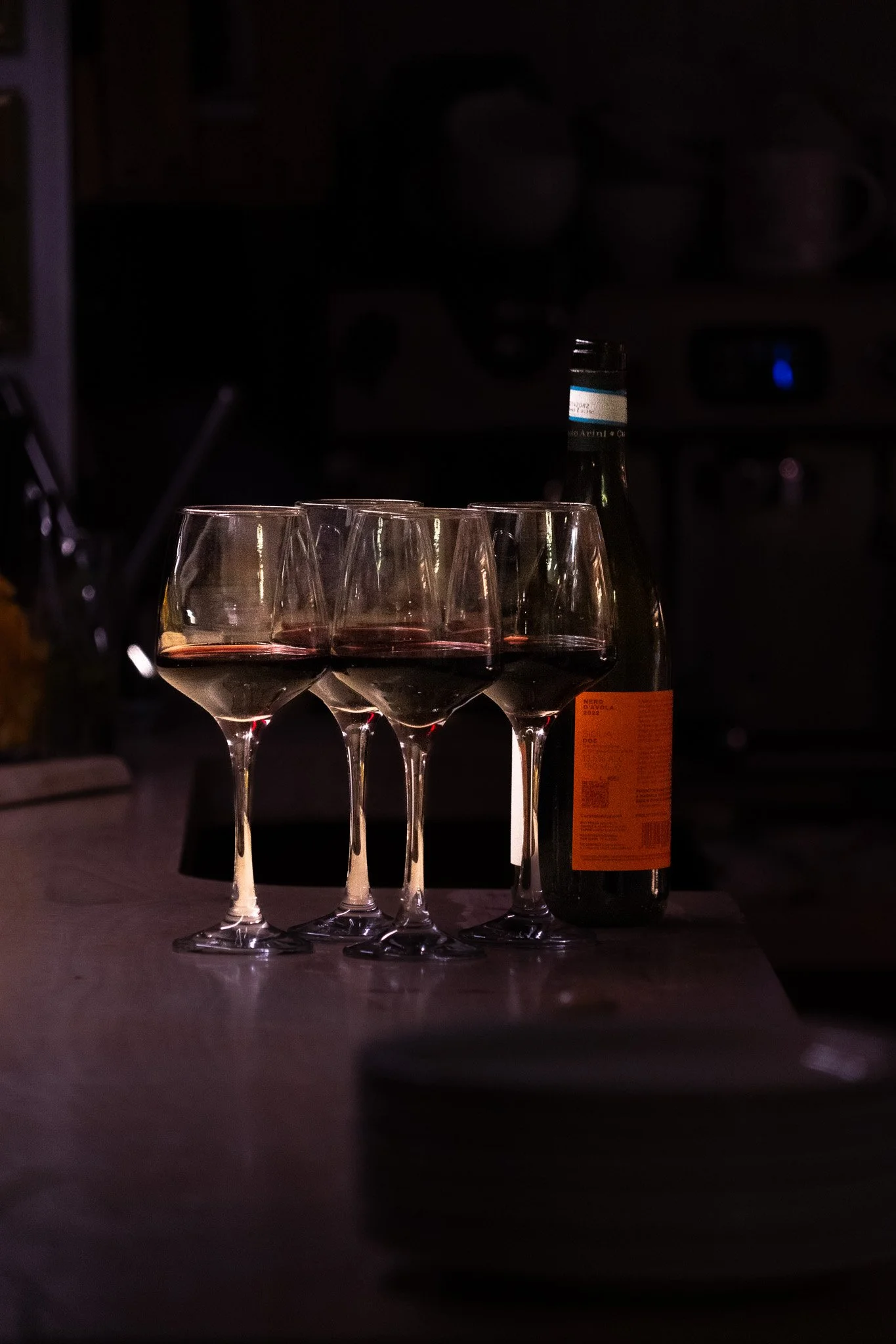Four glasses of red wine and one bottle on a kitchen counter in a dimly lit room.