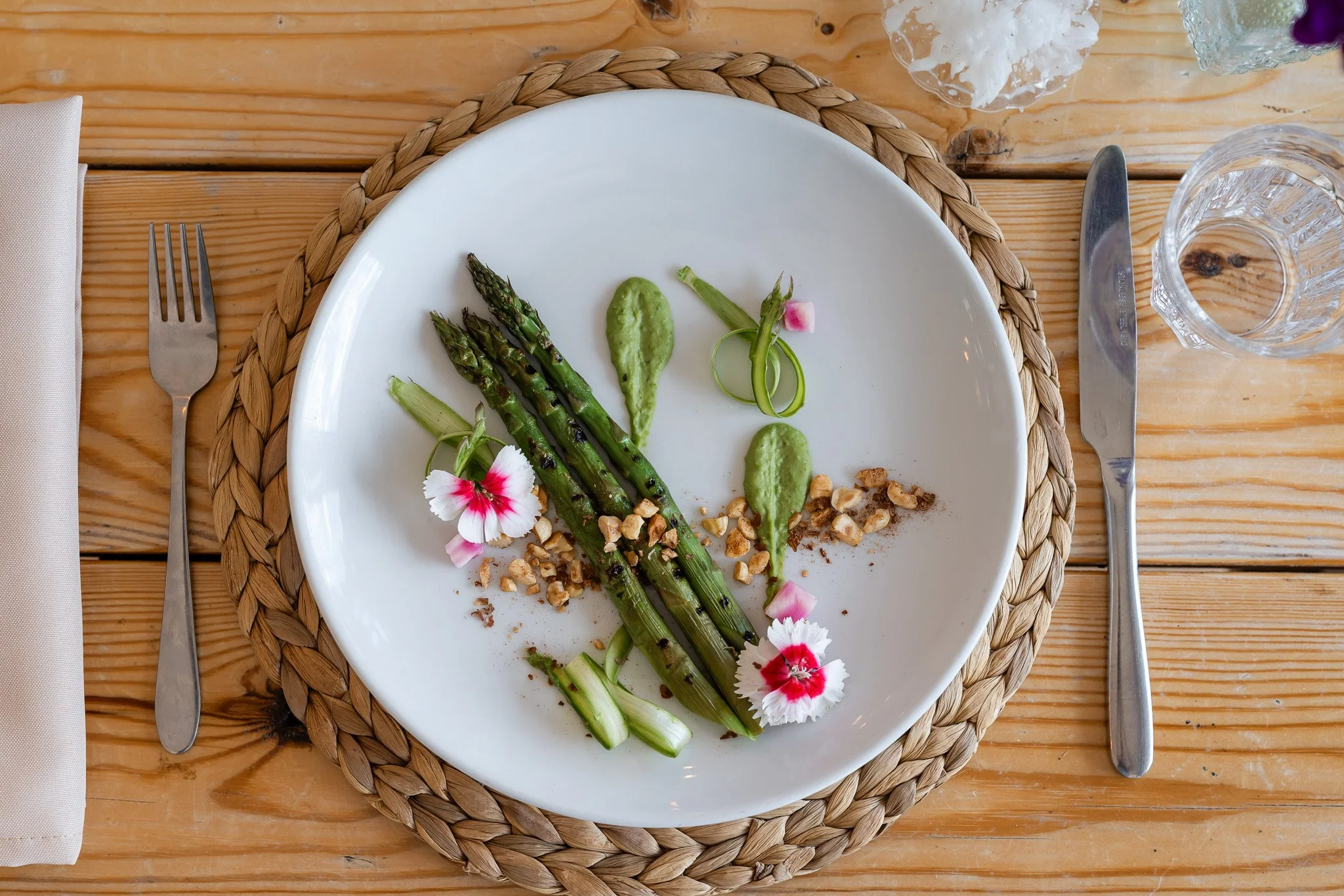 A white plate with grilled asparagus, green sauces, chopped nuts, and edible flowers on a woven placemat, set on a wooden table with a fork on the left and a knife on the right.