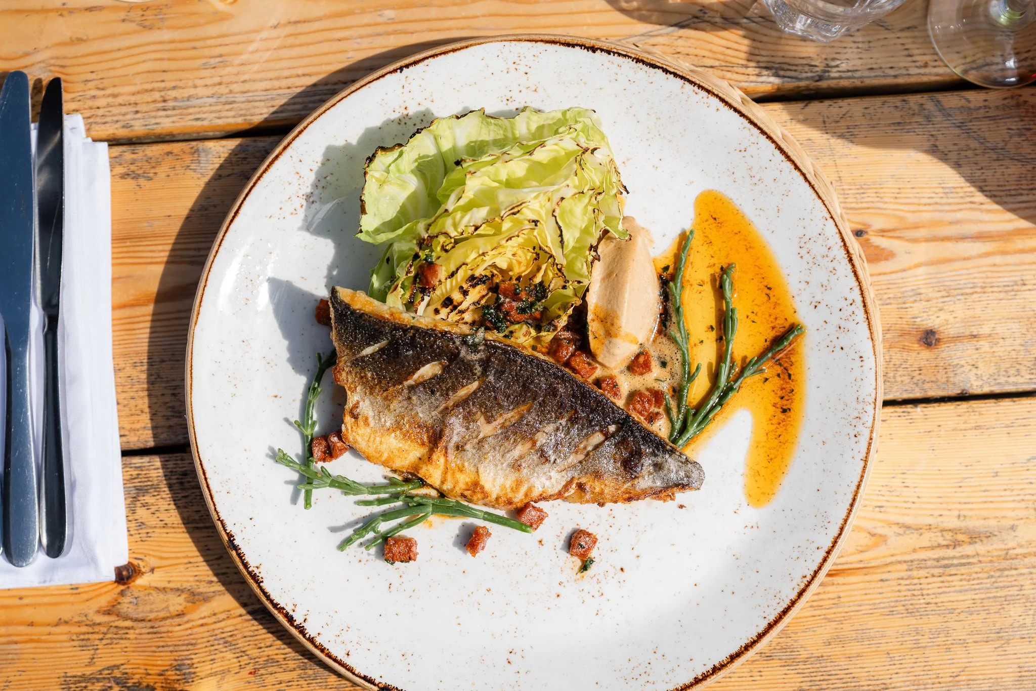 A plate of grilled fish with grilled vegetables, lettuce, and sauce on a wooden table.