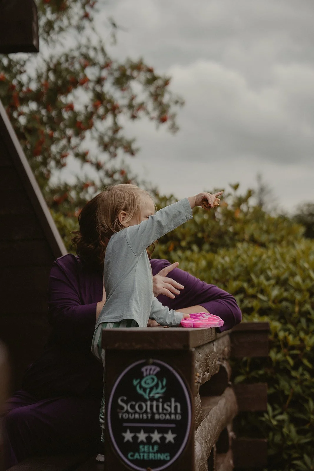 A young girl with light hair pointing at something while standing on a Leny Estate Lodge, with an adult nearby. They are on a wooden deck or balcony with a "Scottish Tourist Board Self Catering" sign visible and the sky is overcast.