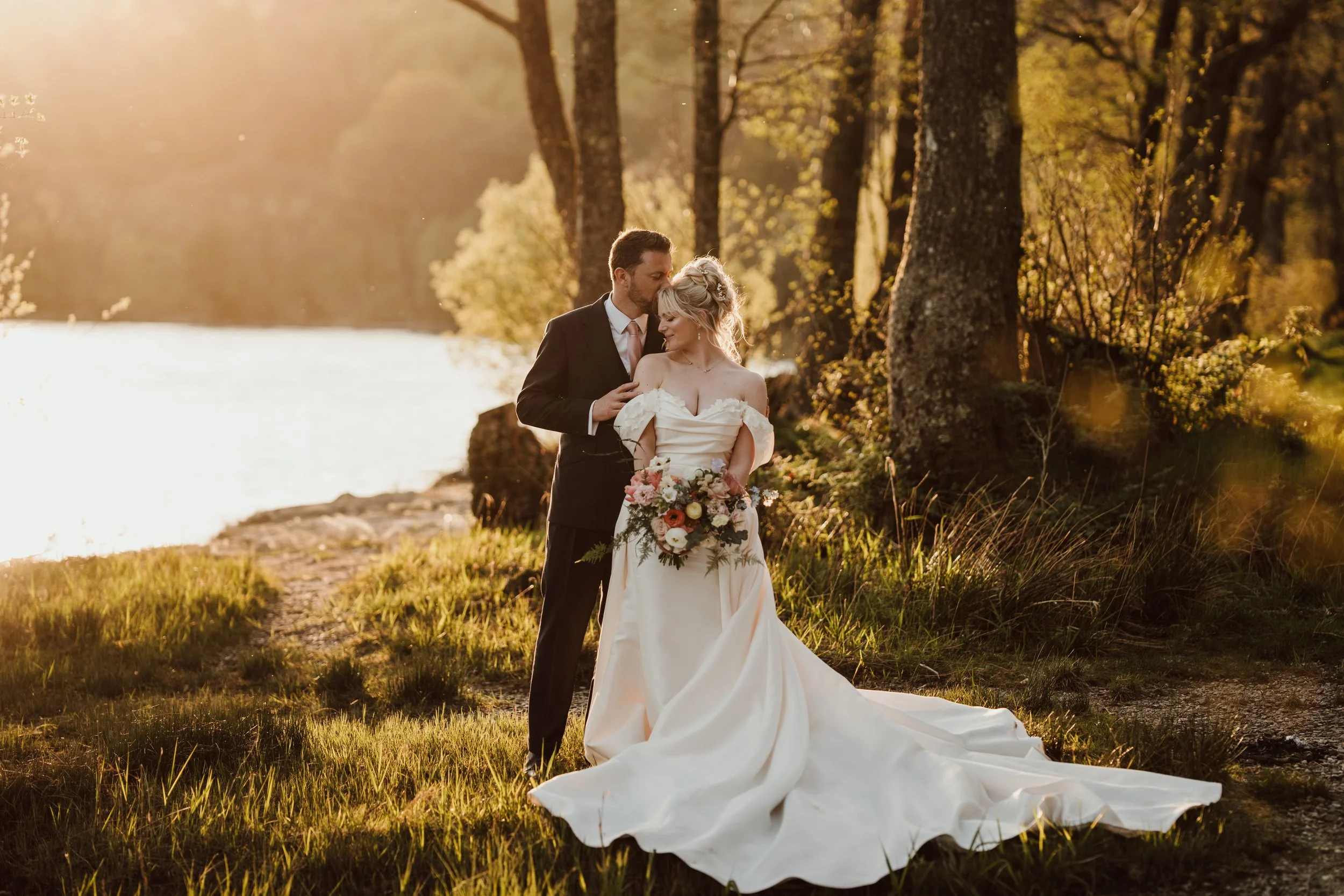 Bride and groom in wedding attire standing by loch Venachar, embracing each other at sunset with trees in the background.