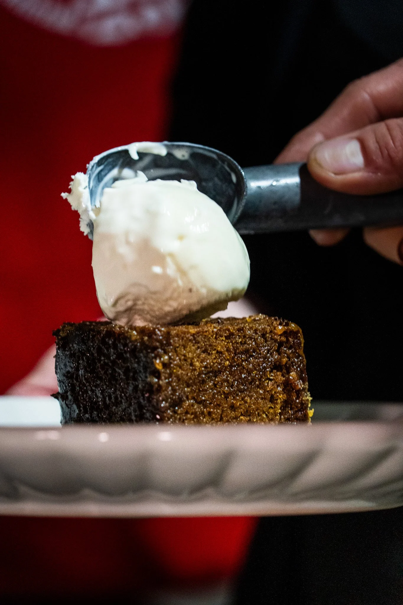 A person holds a spoonful of vanilla ice cream over a slice of chocolate cake on a white plate.