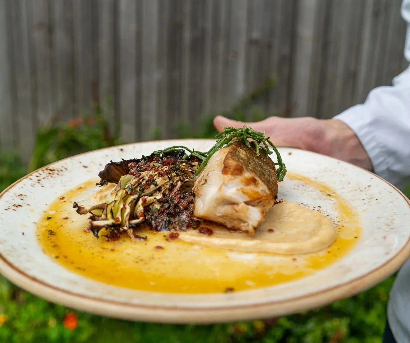 Plate of gourmet dish with cooked fish, roasted vegetables, and sauce, garnished with herbs held by the chef at Venachar Lochside