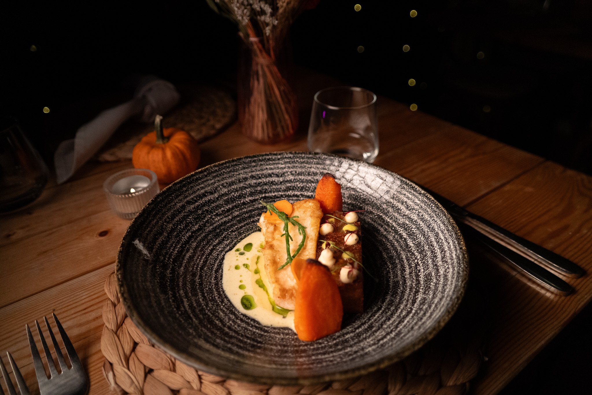 A gourmet dish served on a black textured plate, featuring a piece of fish with garnish, microgreens, roasted carrots, and cream sauce with green oil drizzle, on a wooden table with a glass of water and decorative items in the background.