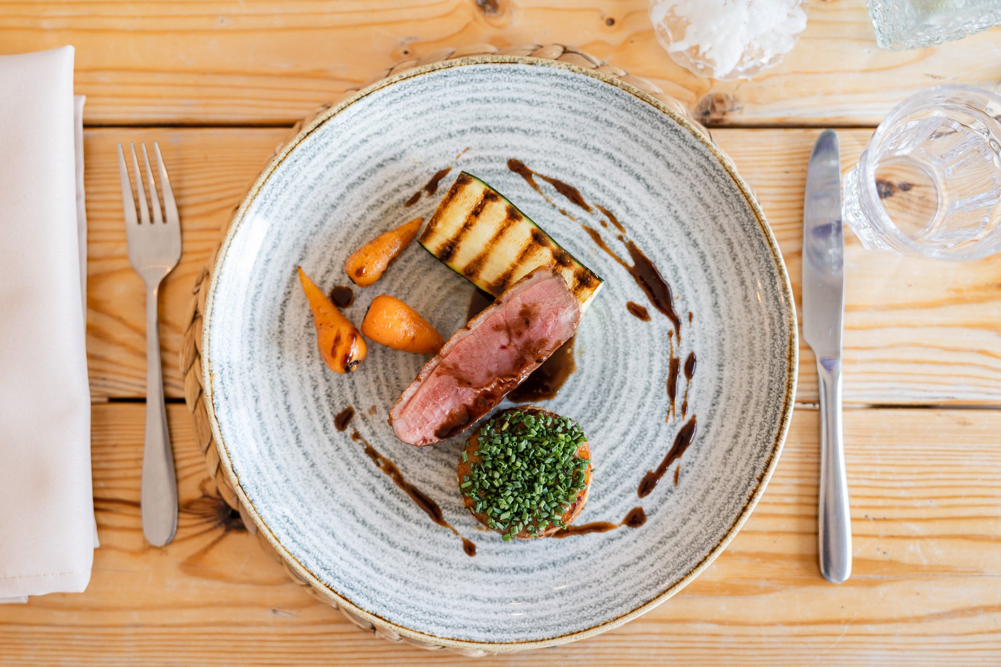 A plated gourmet meal with a grilled venison entrée, grilled zucchini, carrots, and chives, garnished with a dark sauce on a ceramic plate, with a fork and knife on a wooden table.