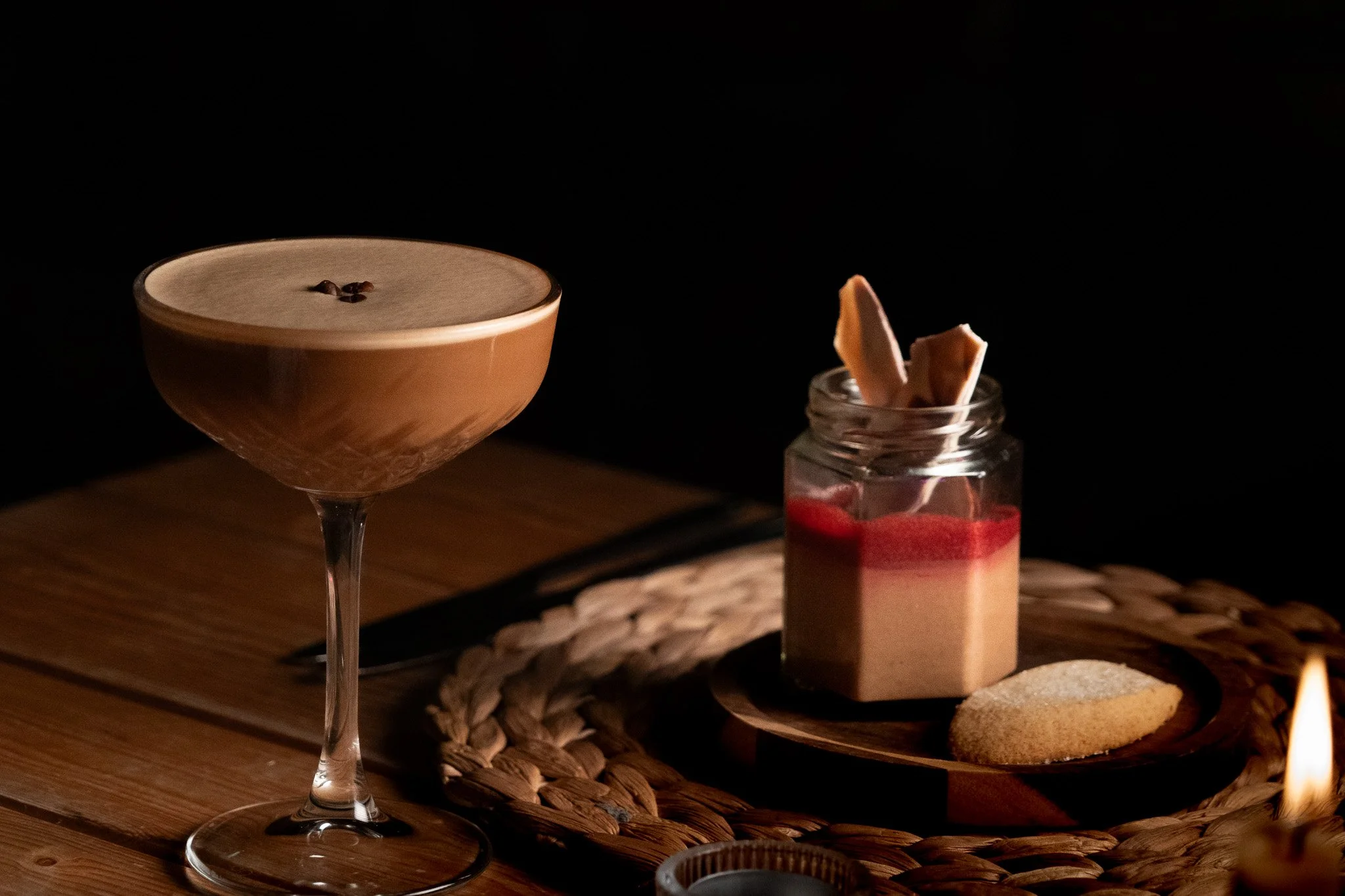 A espresso martini cocktail with coffee beans on top in a coupe glass, alongside a jar of layered dessert with strawberries and cookies, on a wooden table with a woven placemat, illuminated by warm lighting.