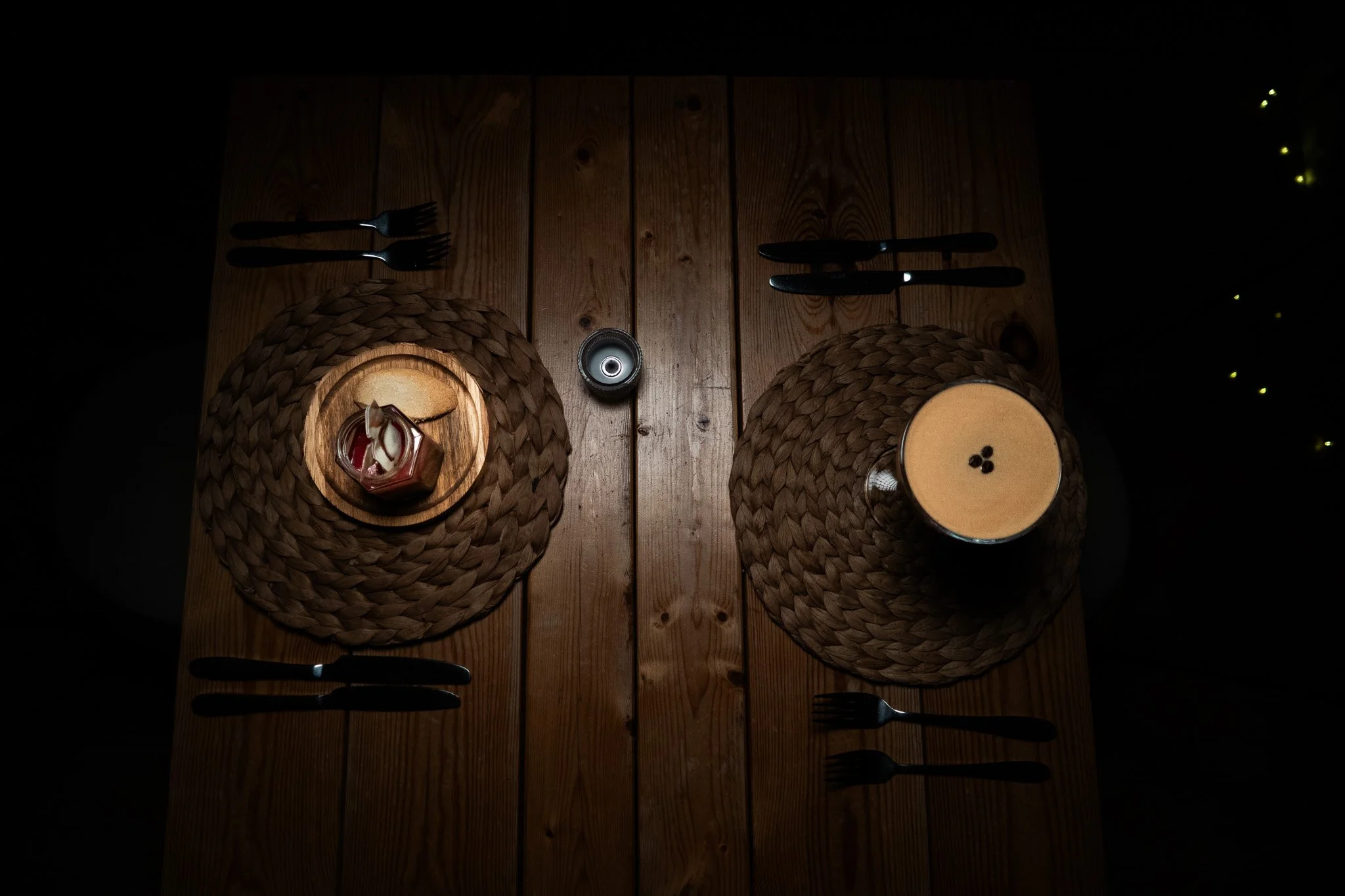 A wooden dining table set for two, with woven placemats, black cutlery, a small chocolate dessert and a espresso martini, viewed from above in dim lighting.