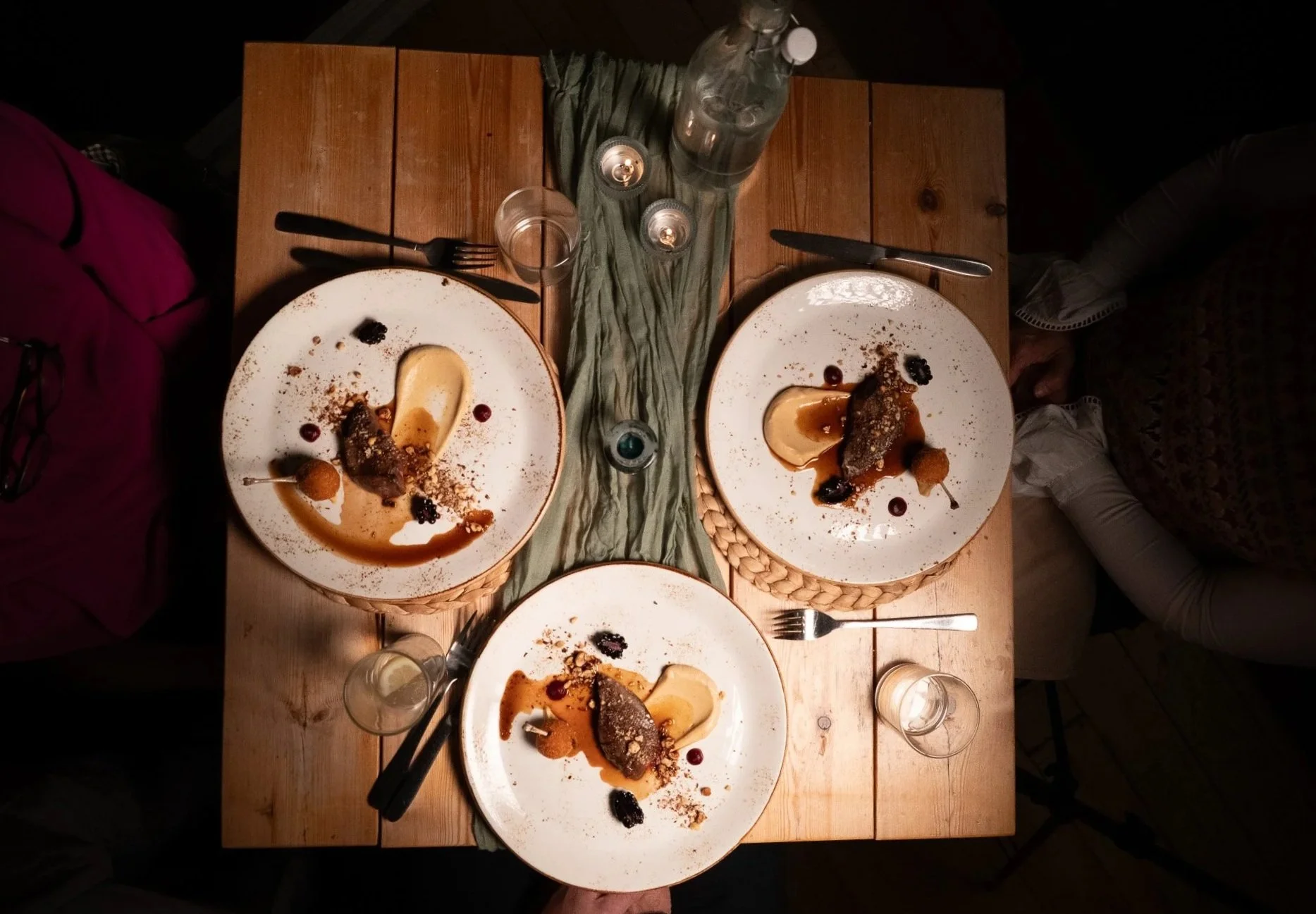 Three dinner plates with wood pigeon, sauce drizzles, and garnishes on a wooden table, surrounded by glasses, utensils, and candles.