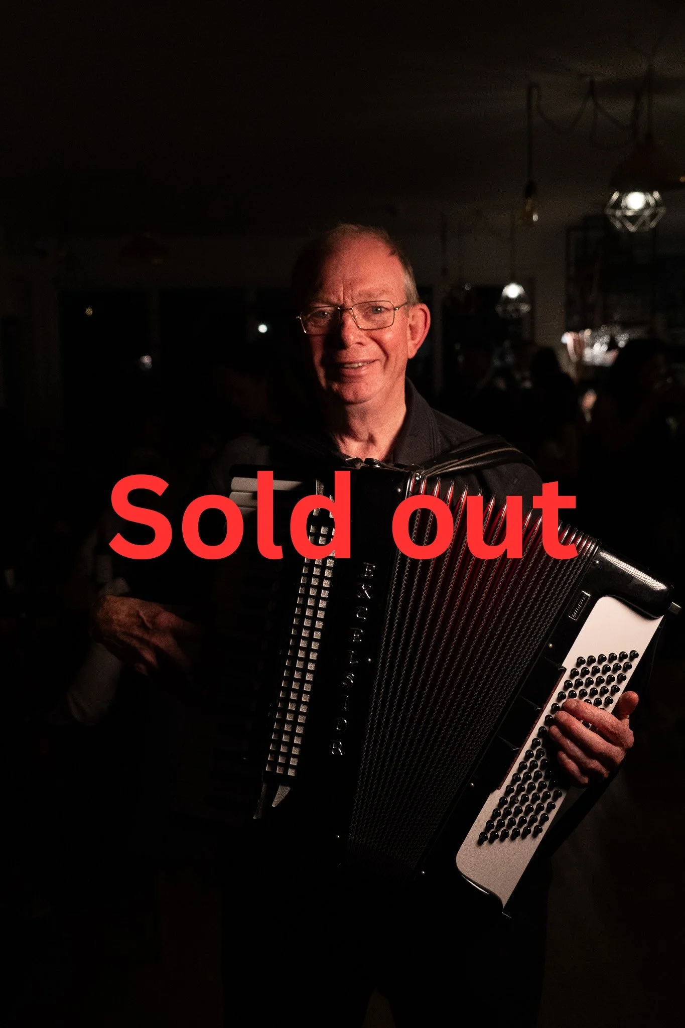 A man holding an accordion at a dark indoor event, with the words 'sold out' overlayed in red.