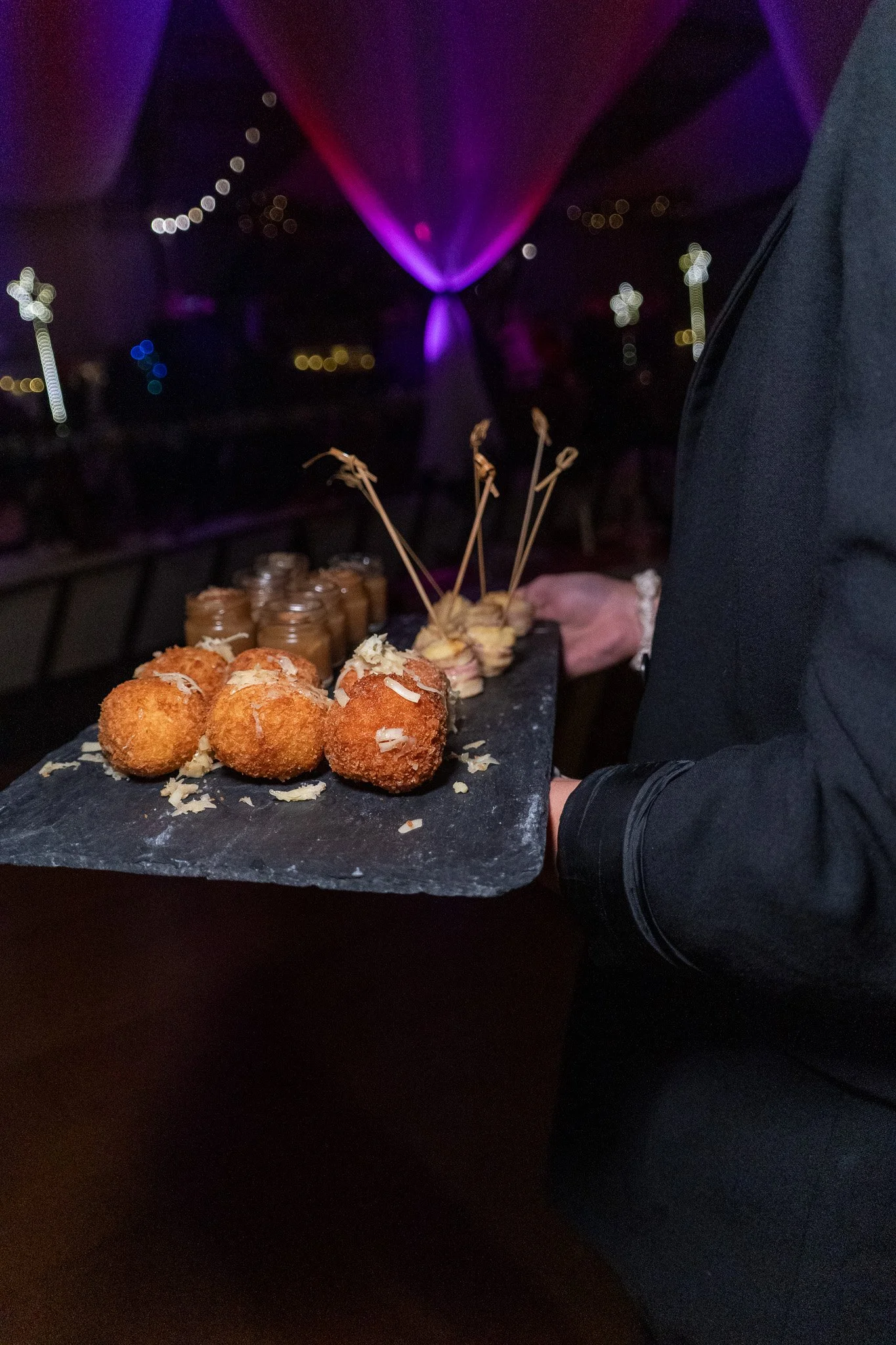 A person holding a tray of assorted canopies, including fried bites and small sandwiches, in a dimly lit setting with purple and blue lighting and blurred background.