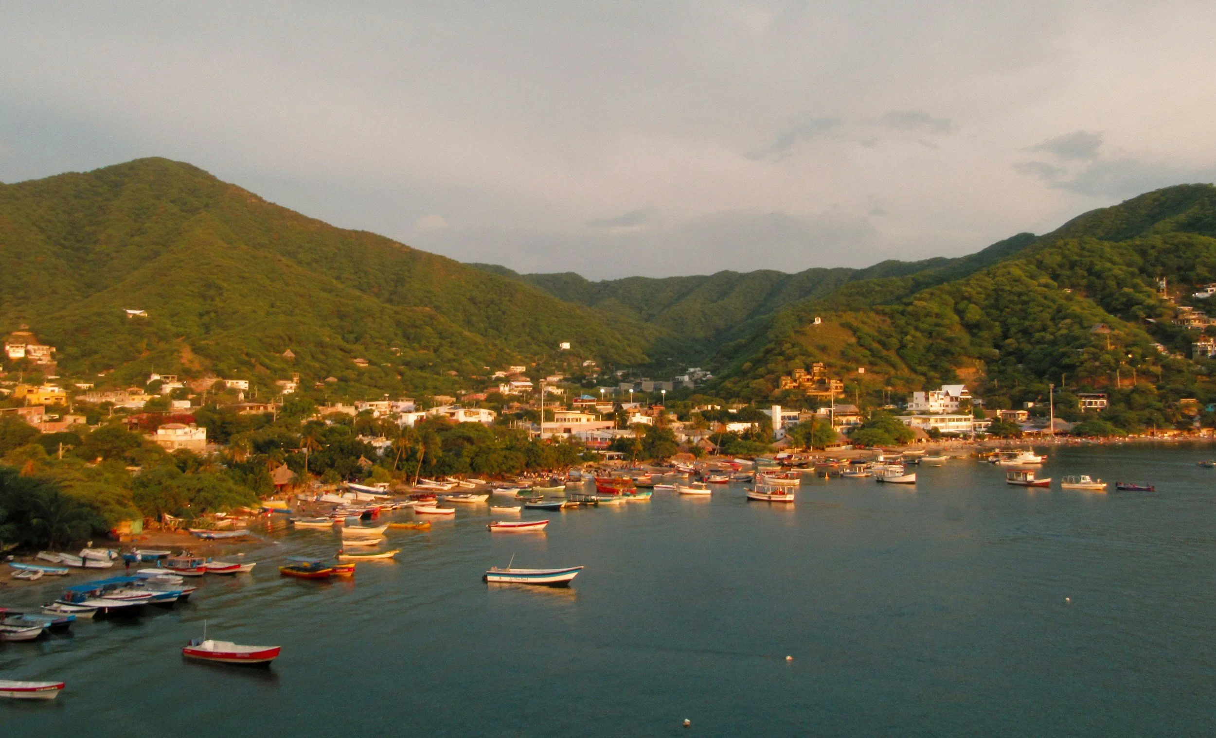 A coastal town with numerous boats docked in the water, surrounded by green hills and mountains under a cloudy sky.