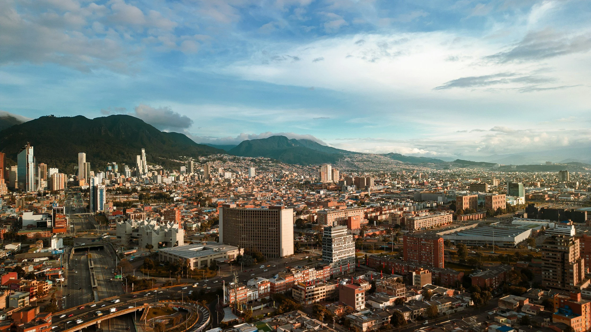 A panoramic view of a cityscape with skyscrapers, residential buildings, and streets, with mountains in the background and a partly cloudy sky overhead.