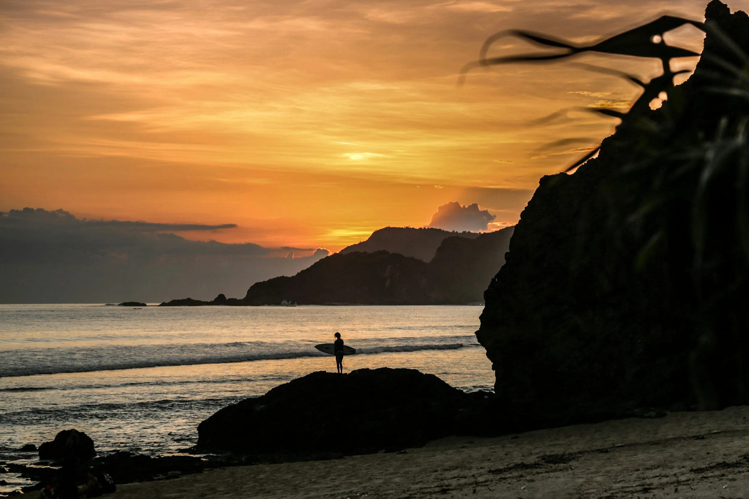 Silhouette of a person holding a surfboard on a beach during sunset, with mountains and a cloudy sky in the background.