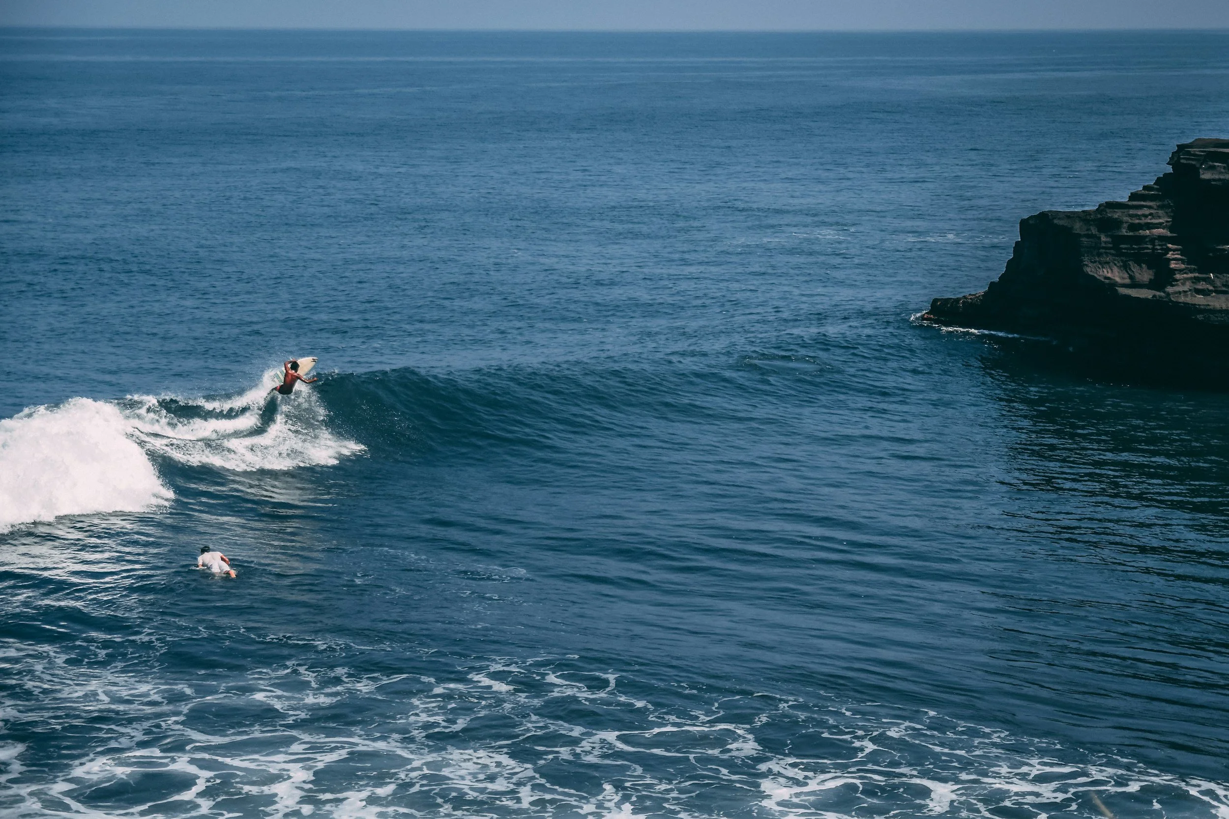 A person surfing on a wave near a rocky cliff in the ocean.