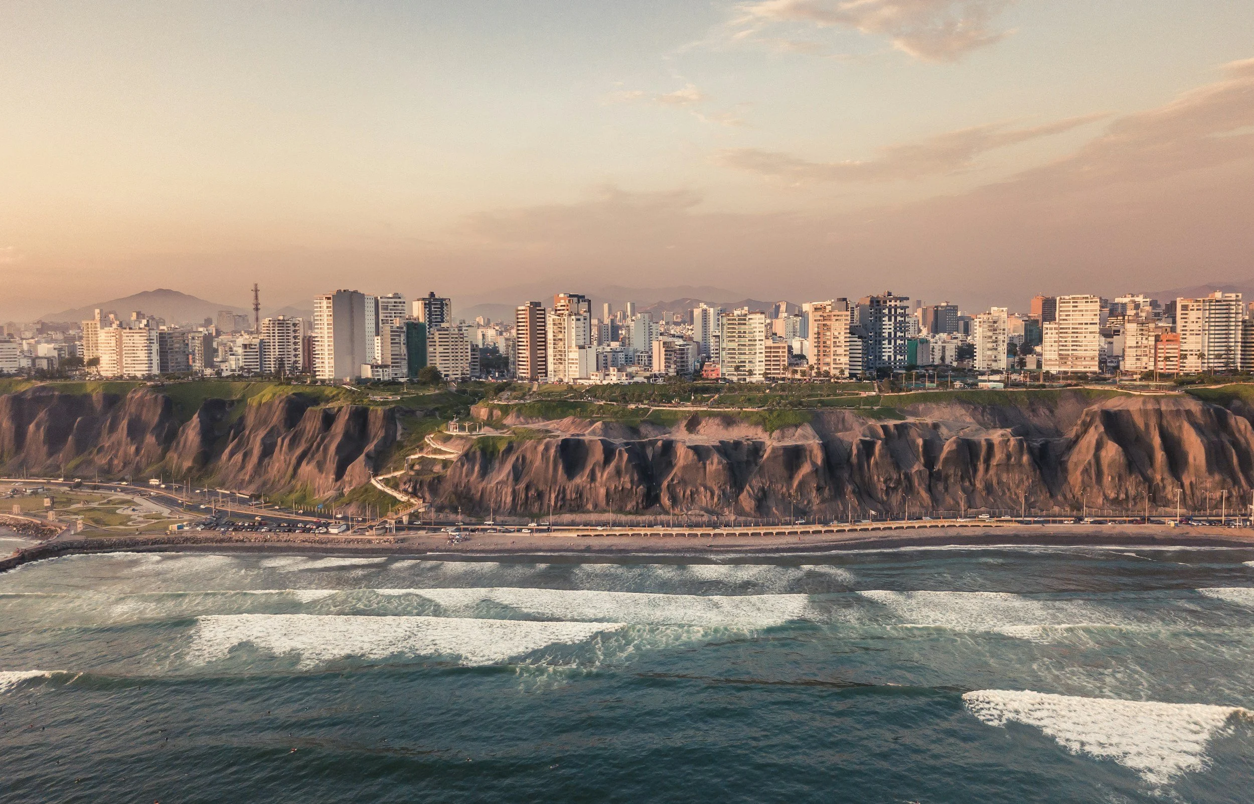 City skyline with numerous high-rise buildings, a beach with waves in the foreground, and a mountain range in the background during sunset or sunrise.