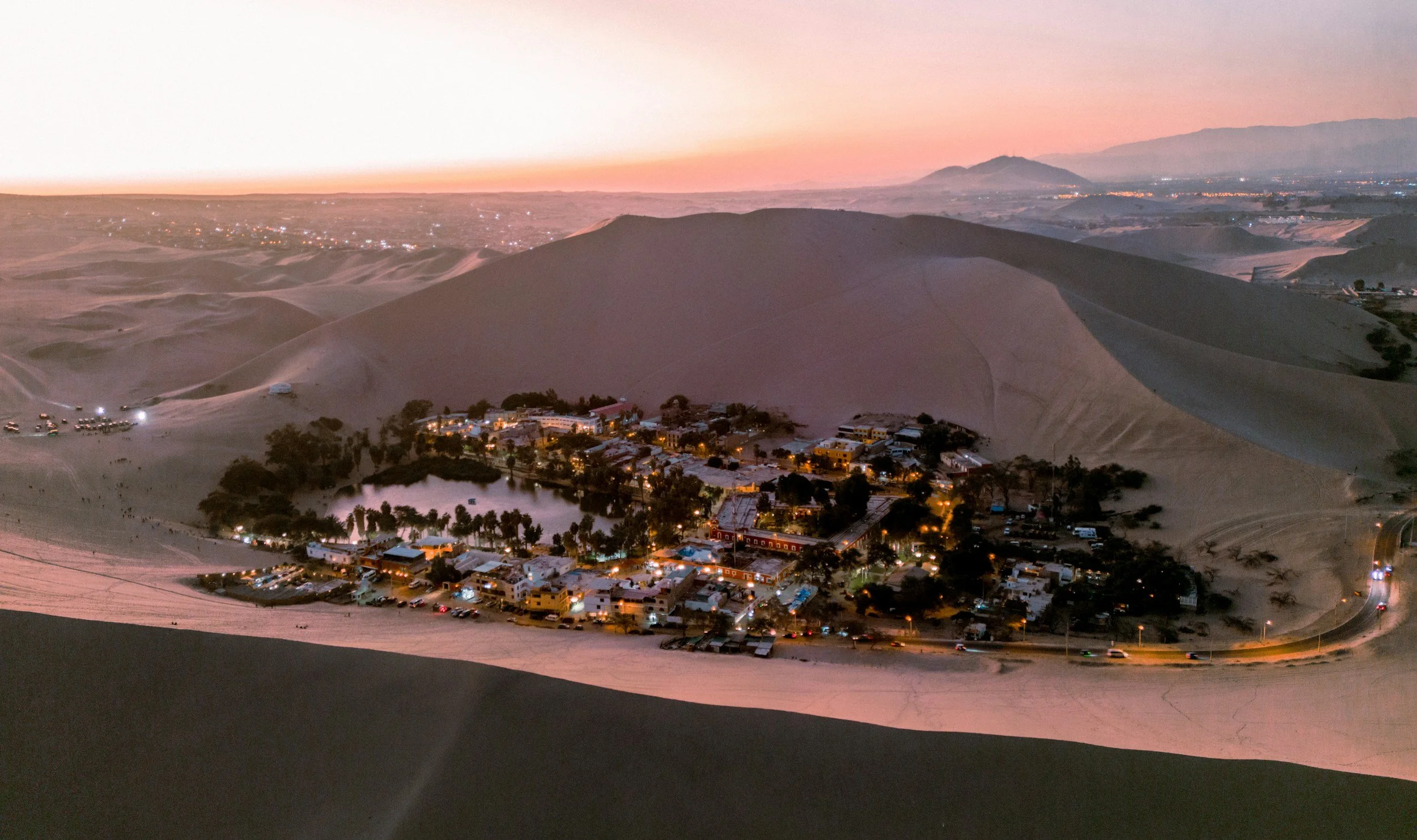 View of sand dunes at sunset with a small village and a pond at the base, surrounded by trees, with mountains in the distance.