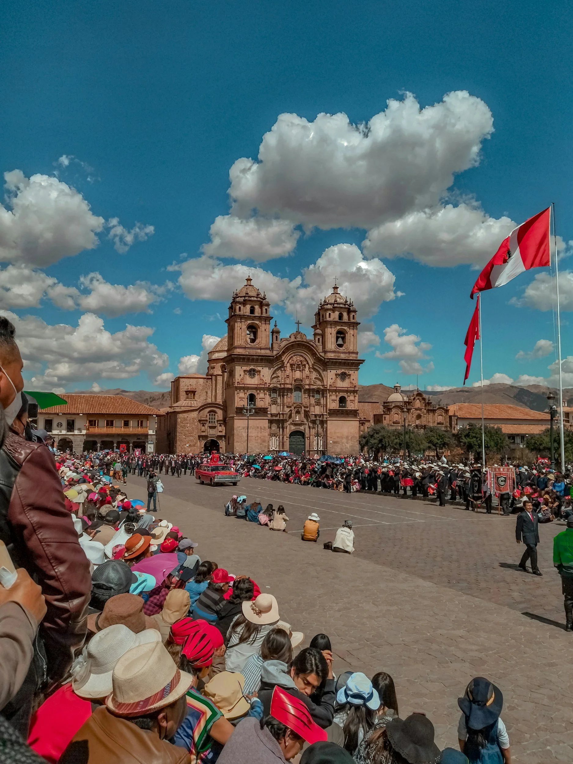 A large crowd of people sitting and standing in a plaza in front of an ornate historic church with twin bell towers, under a blue sky with scattered clouds. Some people are holding umbrellas and wearing hats.