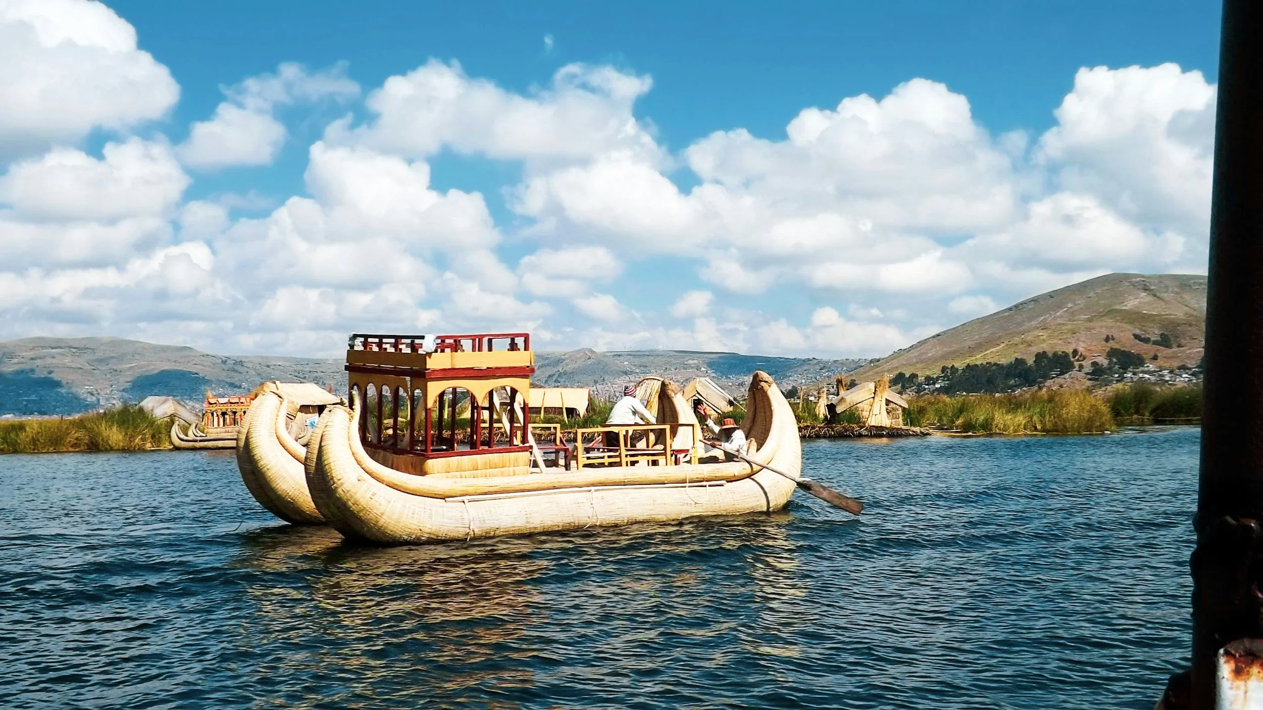 A boat designed like a traditional animal-shaped vessel with a curved body and decorative details floats on a river, with hills and a partly cloudy sky in the background.