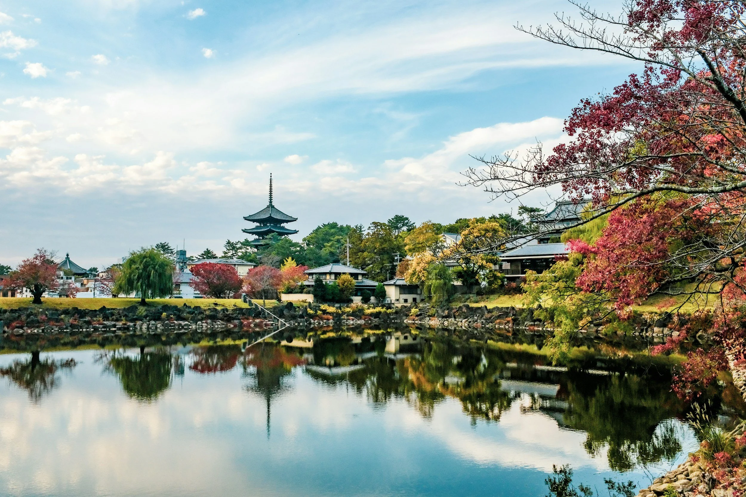 Scenic view of a traditional Japanese temple with multiple stories and a pagoda roof, surrounded by colorful autumn trees, reflecting in a calm body of water.