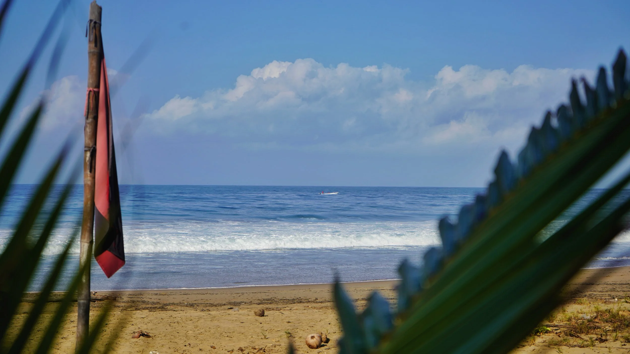 Beach scene with a flag on a pole, sandy shoreline, ocean waves, a boat in the distance, and partly cloudy sky. On the beach at Playa Larga, Puerta Paraiso Boutique Hotel perfect, best wedding destination in Zihuatanejo