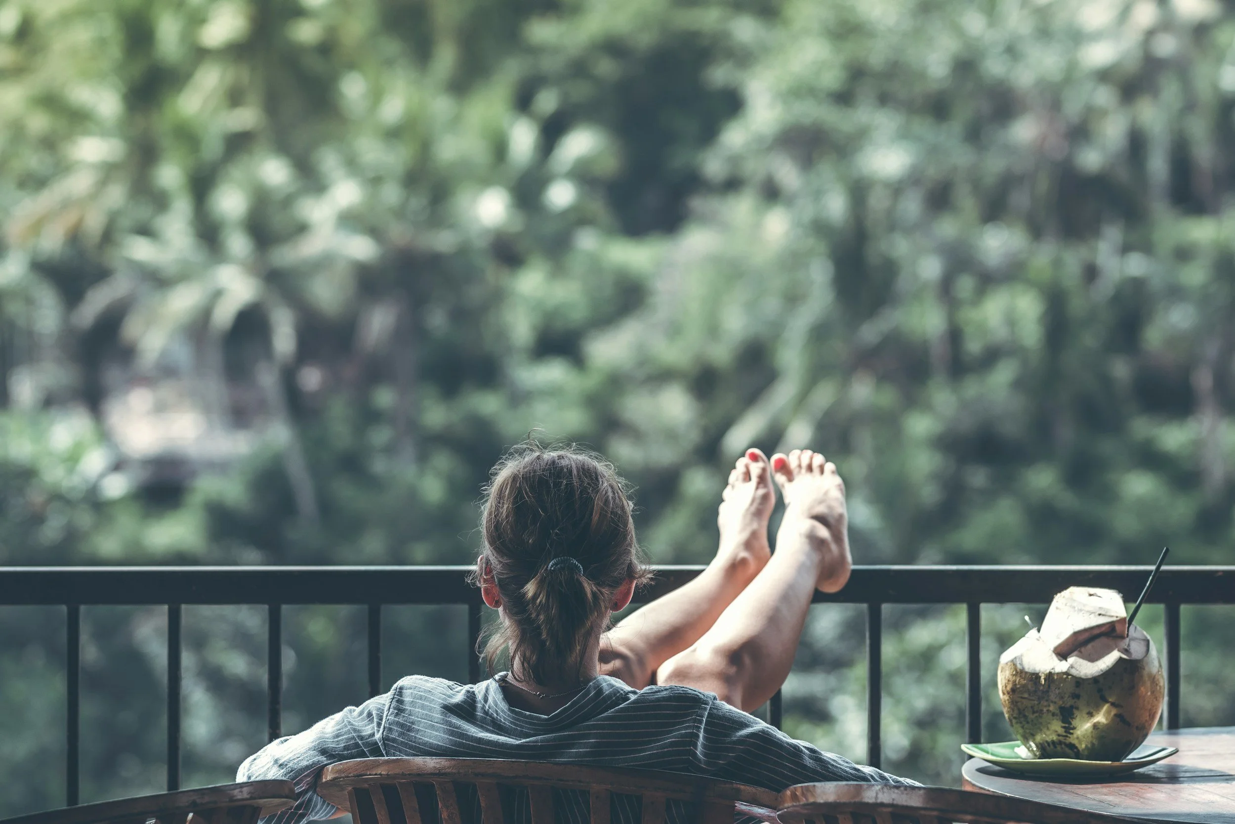 A woman relaxing on a balcony, with her feet resting on the railing, overlooking a lush green forest. A coconut with a straw and a bowl are on a table nearby.