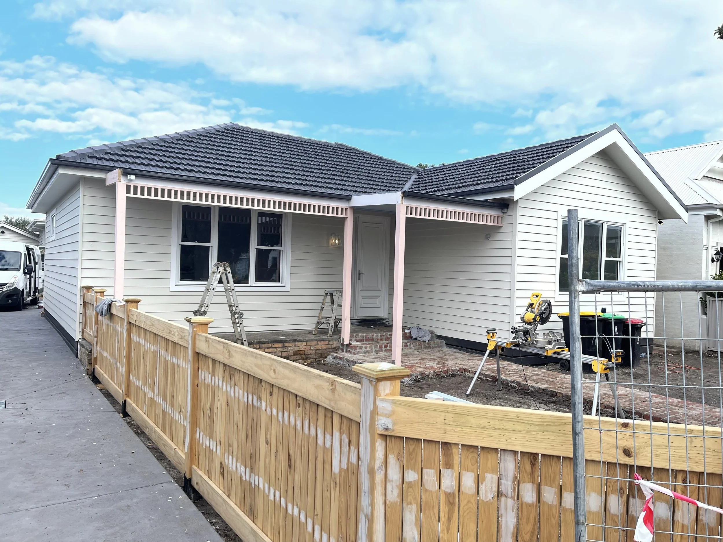 House under construction with a wooden fence, scaffolding, tools, and brickwork, white siding, black roof, cloudy sky.