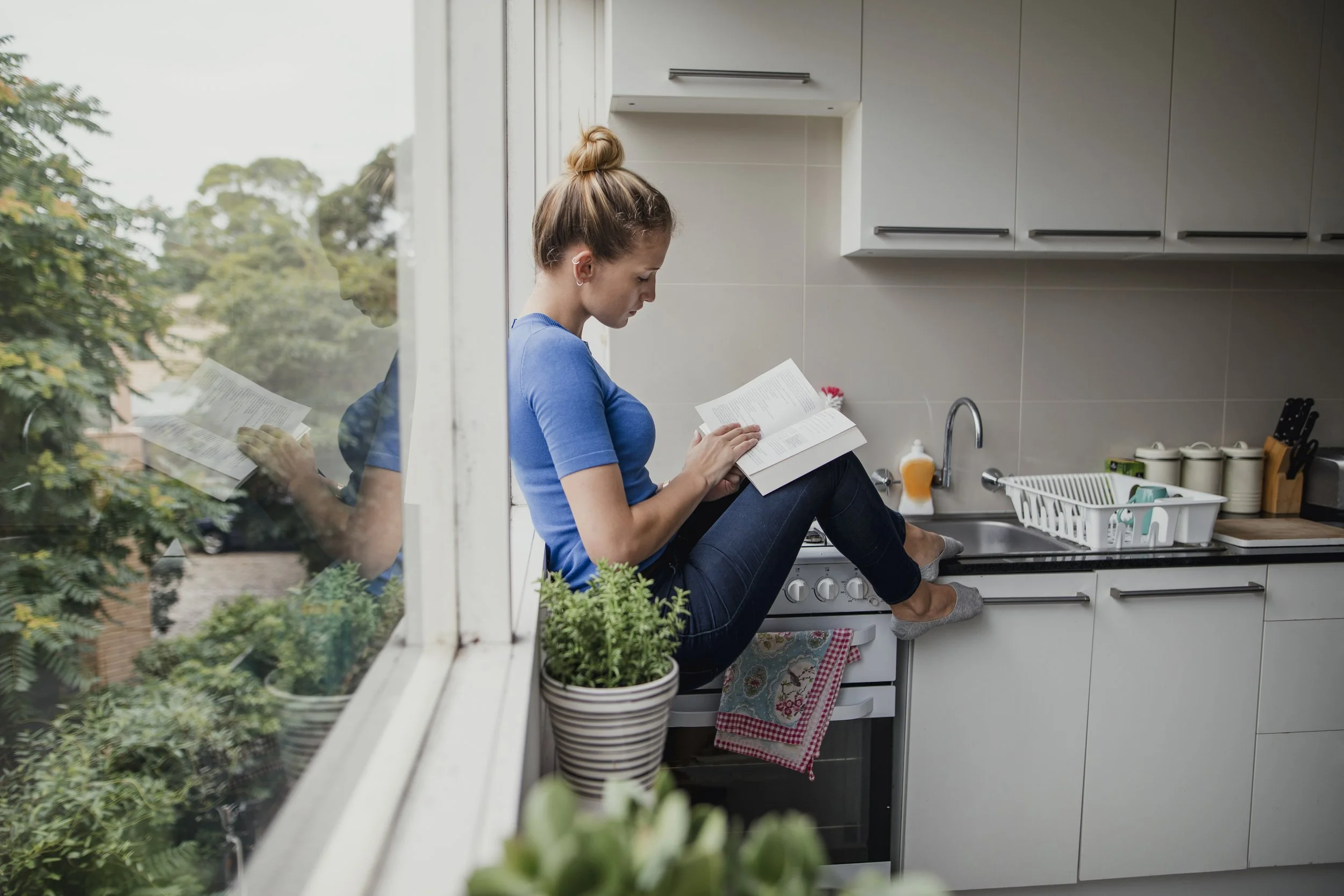A woman with red hair tied in a bun, wearing a blue shirt and gray socks, sitting on a kitchen stove with her legs crossed, reading a book. She is next to a window with a view of trees outside. The kitchen has white cabinets, a sink, dish rack, and kitchen utensils.