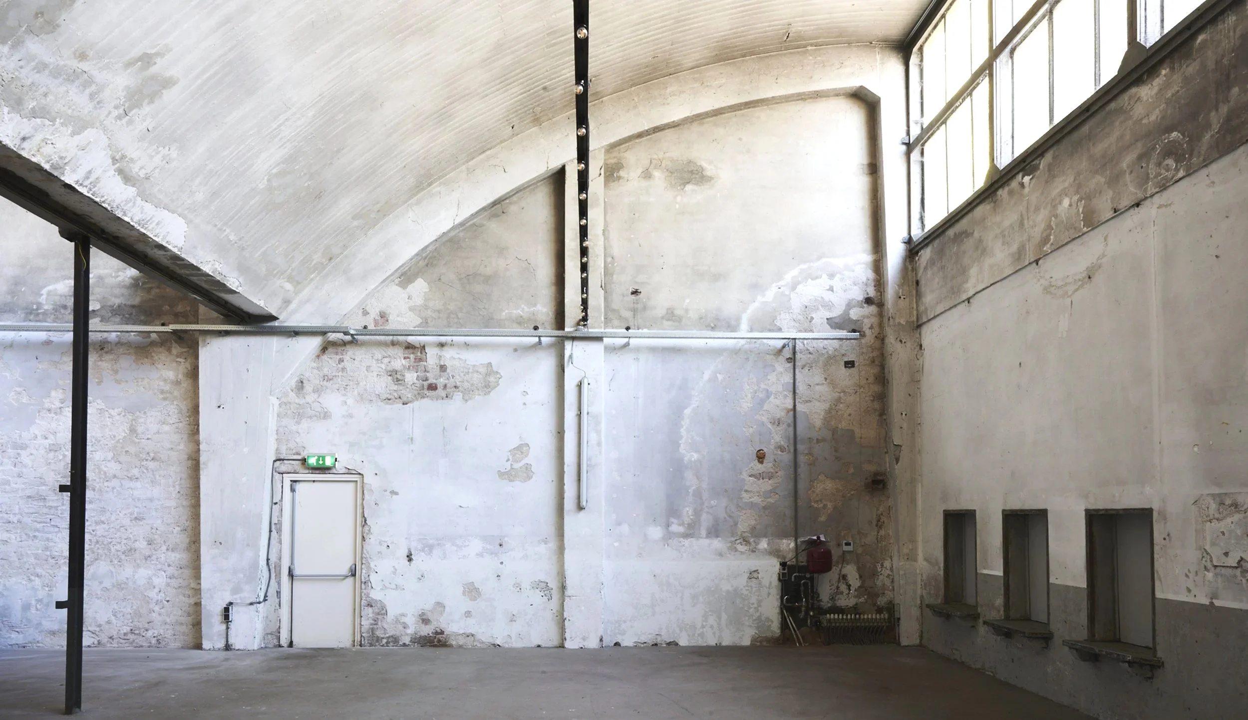 Interior of an industrial or warehouse space with high arched ceiling, weathered white walls, three small rectangular windows, and a white door with an illuminated exit sign. Exposed pipes and electrical conduits are visible | Rental Studio 