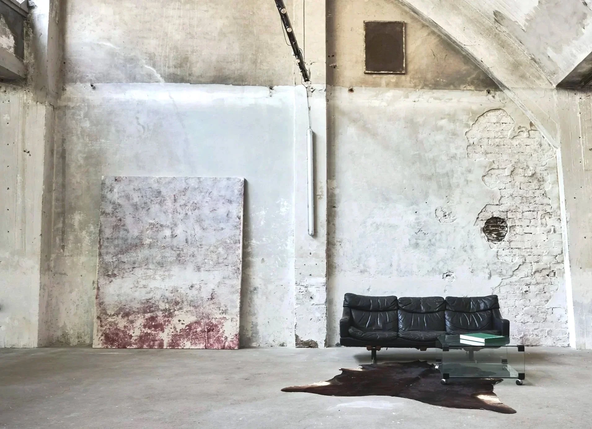 Industrial-style room with exposed brick and concrete walls, a black leather couch, a glass coffee table, a cowhide rug, and a large canvas leaning against the wall | Rental Studio for Film, Photography and Events | Hembrug Zaandam | Greater Amsterda
