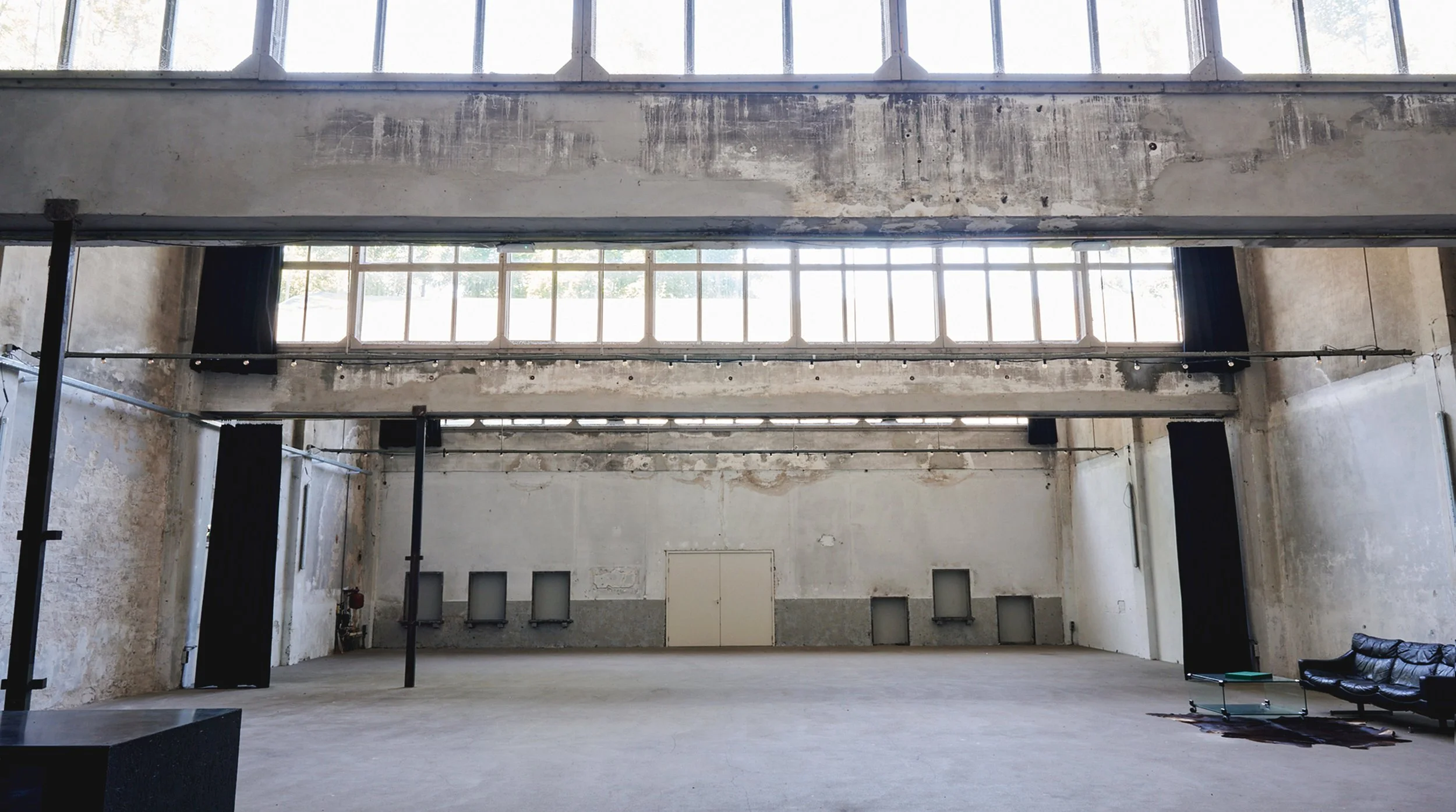 An empty, industrial-style interior with high ceilings, large windows, exposed concrete walls, black curtains, and minimal furniture | Rental Studio for Film, Photography and Events | Hembrug Zaandam | Greater Amsterdam