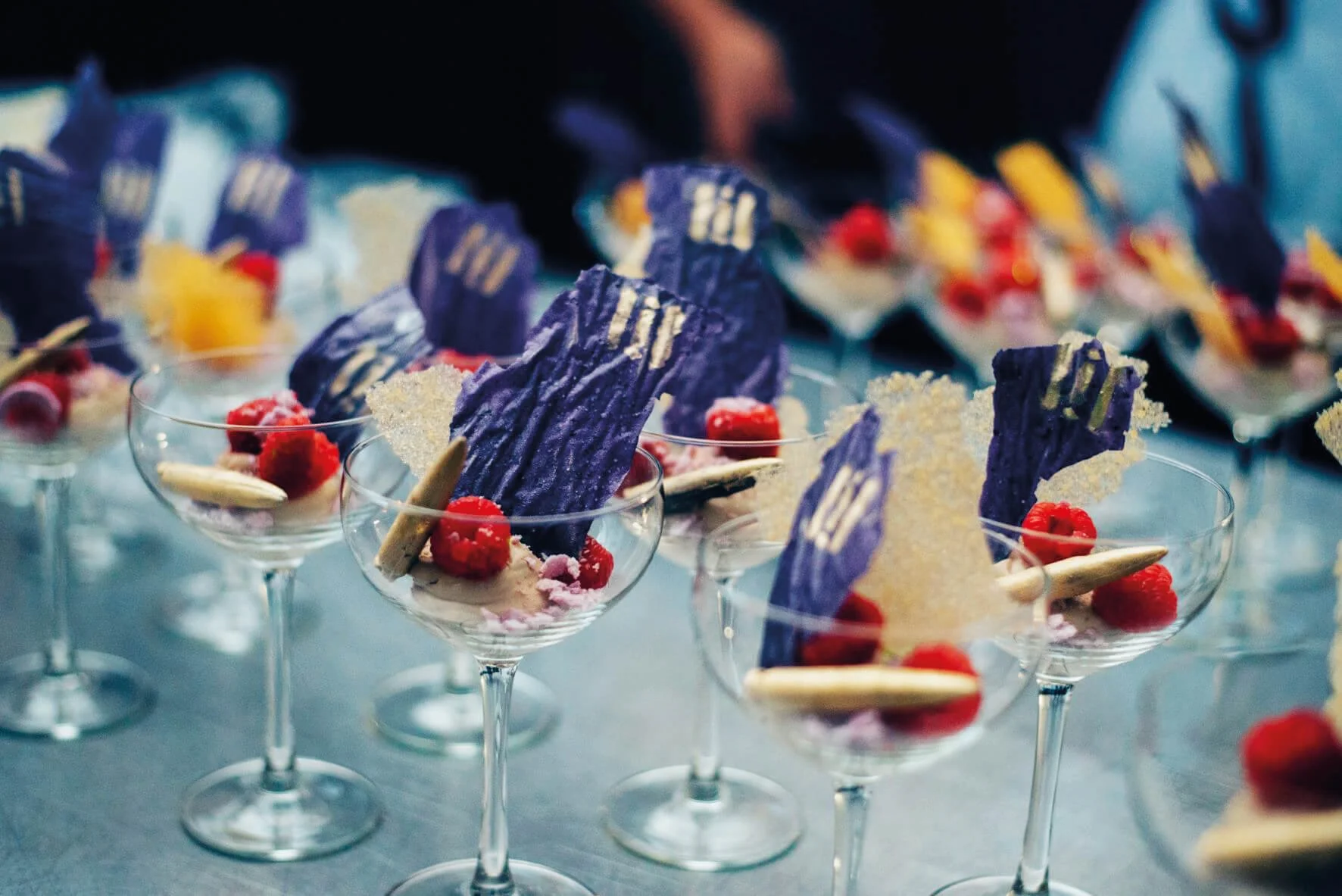 Multiple elegant dessert glasses filled with colorful pastries, raspberries, and purple and yellow decorative toppings on a grey surface | Rental Studio for Film, Photography and Events